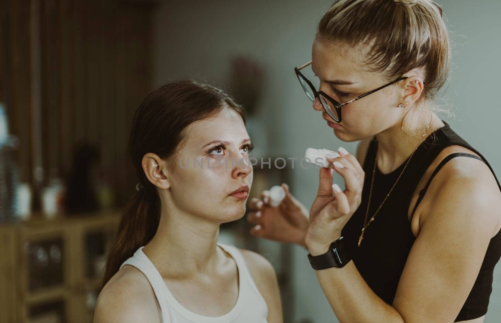 One young handsome Caucasian makeup artist is preparing to apply a moisturizing medicinal liquid under the eyes of a girl sitting in a chair early in the morning in a beauty salon, close-up side view.Step by step.