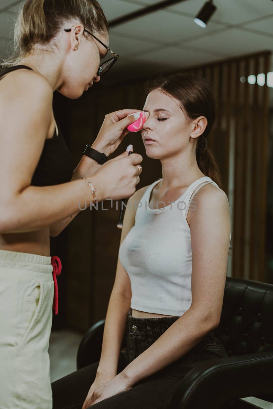 One young handsome Caucasian makeup artist applies foundation with a pink sponge to the eyes of a girl sitting in a chair early in the morning in a beauty salon, close-up side view. Step by step.