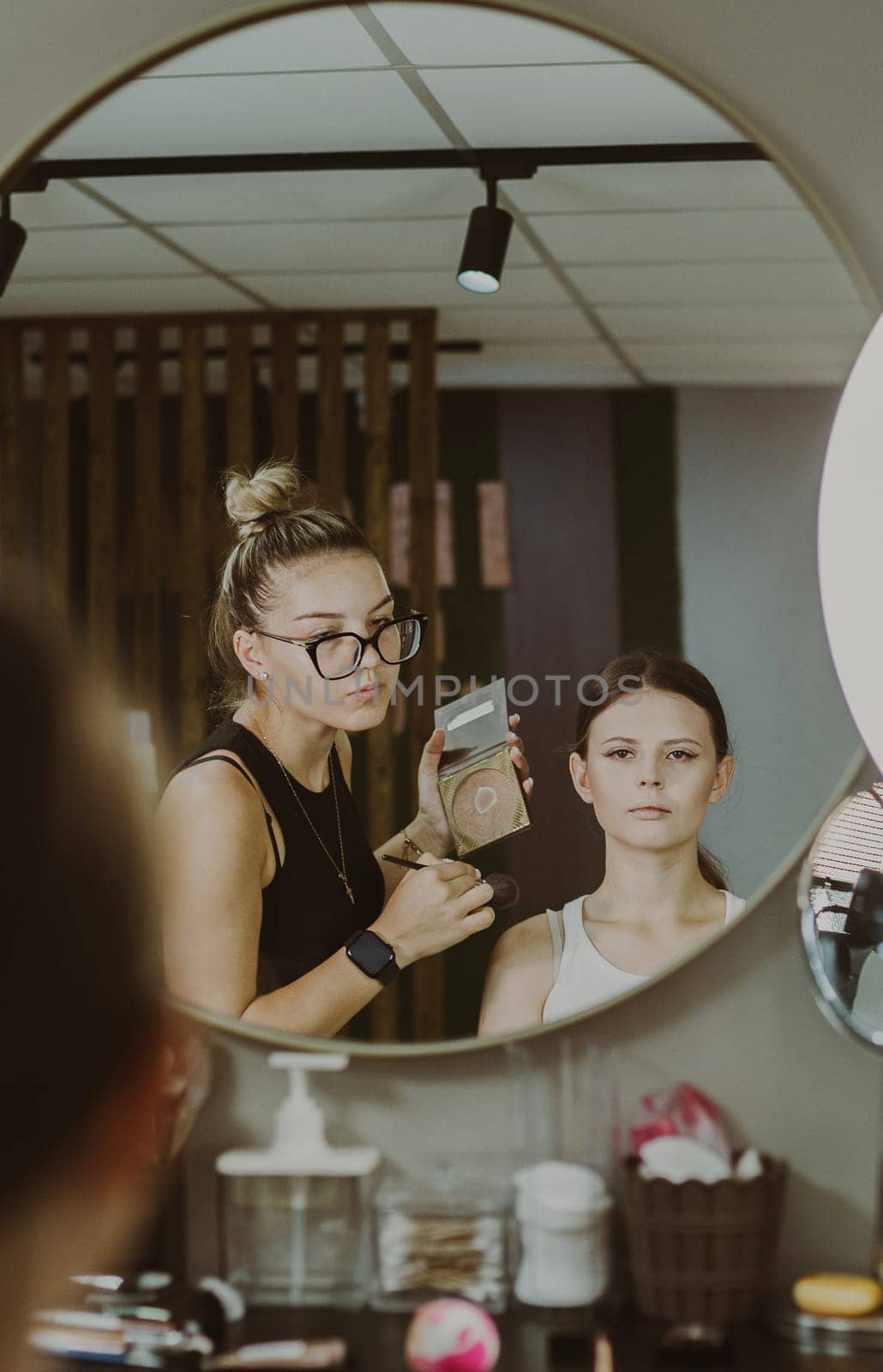 One young beautiful Caucasian makeup artist looks in the mirror at a girl who is doing makeup early in the morning in a beauty salon, close-up side view. Step by step.