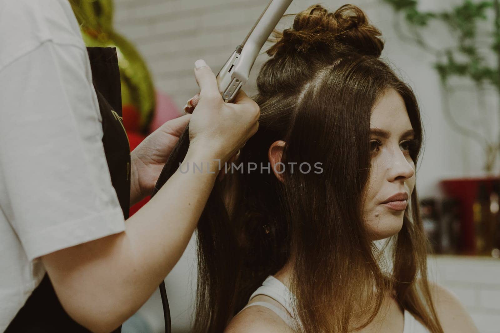 Portrait of a young Caucasian girl sitting sideways in a hairdresser's shop, where the master is curling her hair on a flat iron, close-up view.
