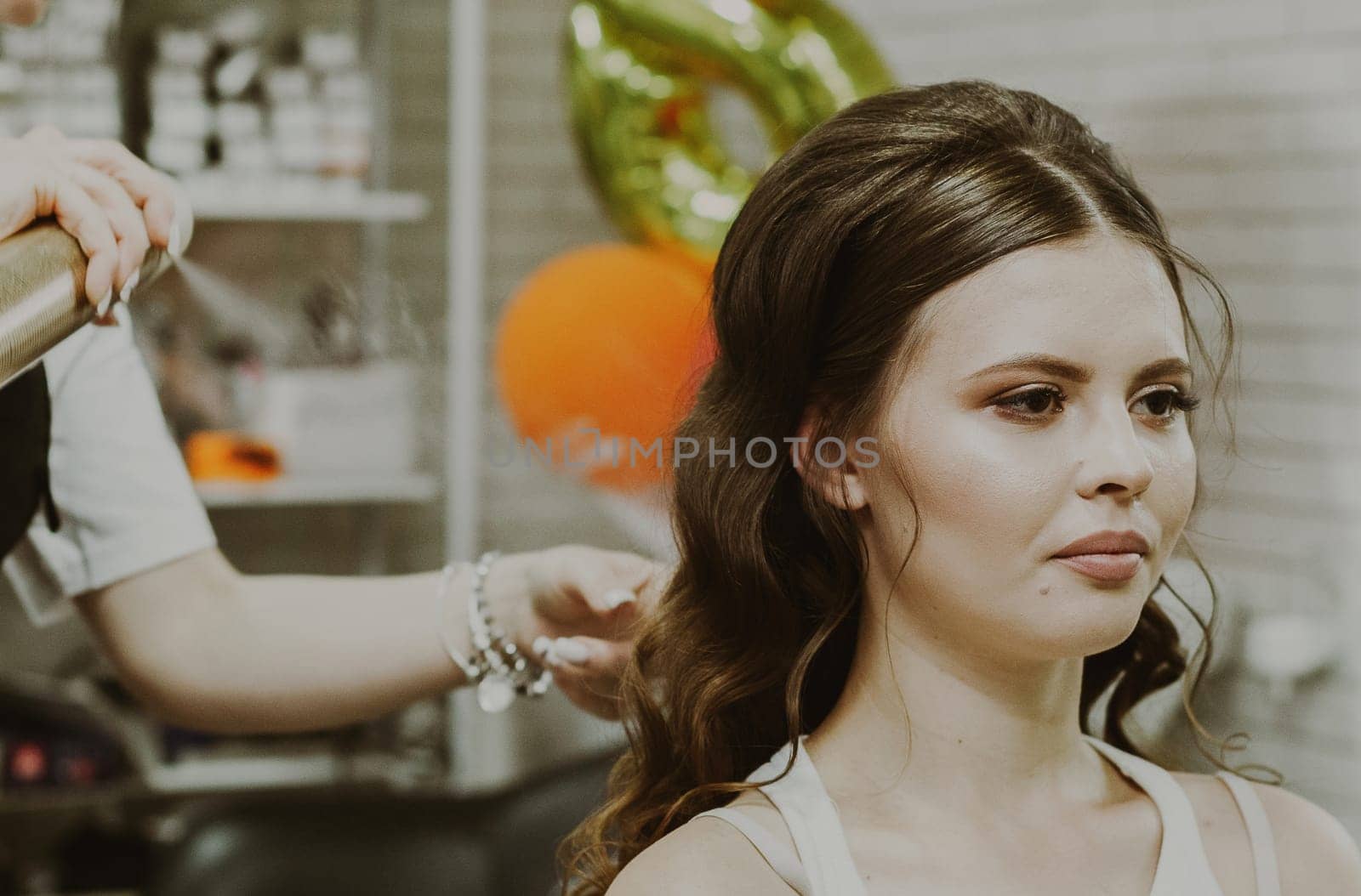 Portrait of a young beautiful Caucasian brunette girl sitting in a hair salon where the master is spraying hairspray on her hair with curls, close-up from the side.