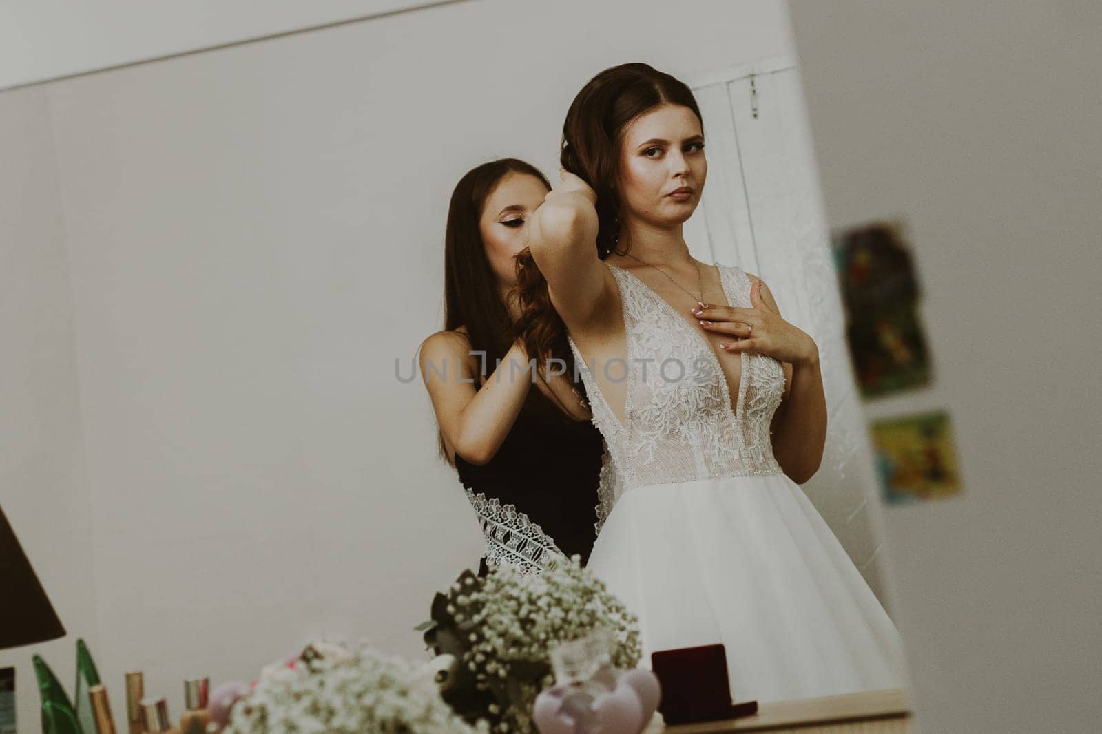 Portrait of a beautiful brunette bride and bridesmaid who fastens the clasp of a necklace around her neck, standing in a room reflected in a mirror, close-up side view.