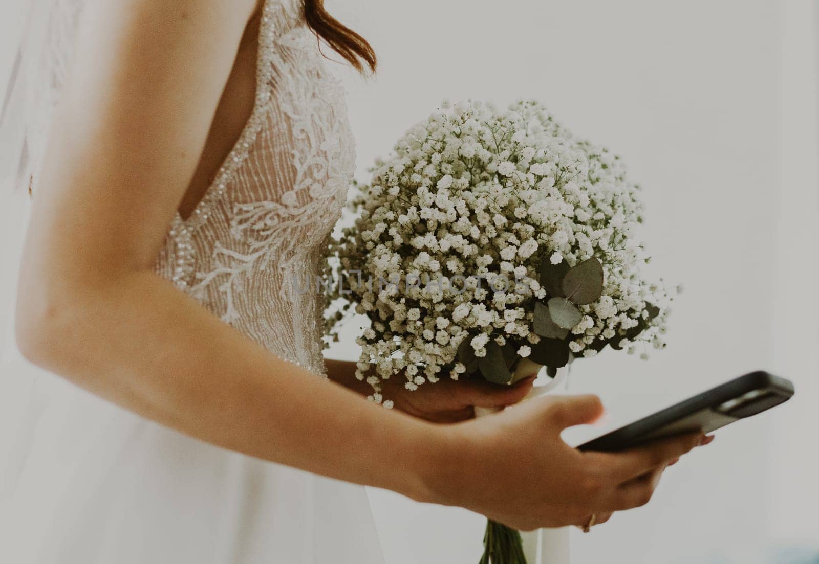 Portrait of one unrecognizable young Caucasian bride standing sideways and holding a bouquet of white boutonnieres and a smartphone in her hands, close-up side view with selective focus.