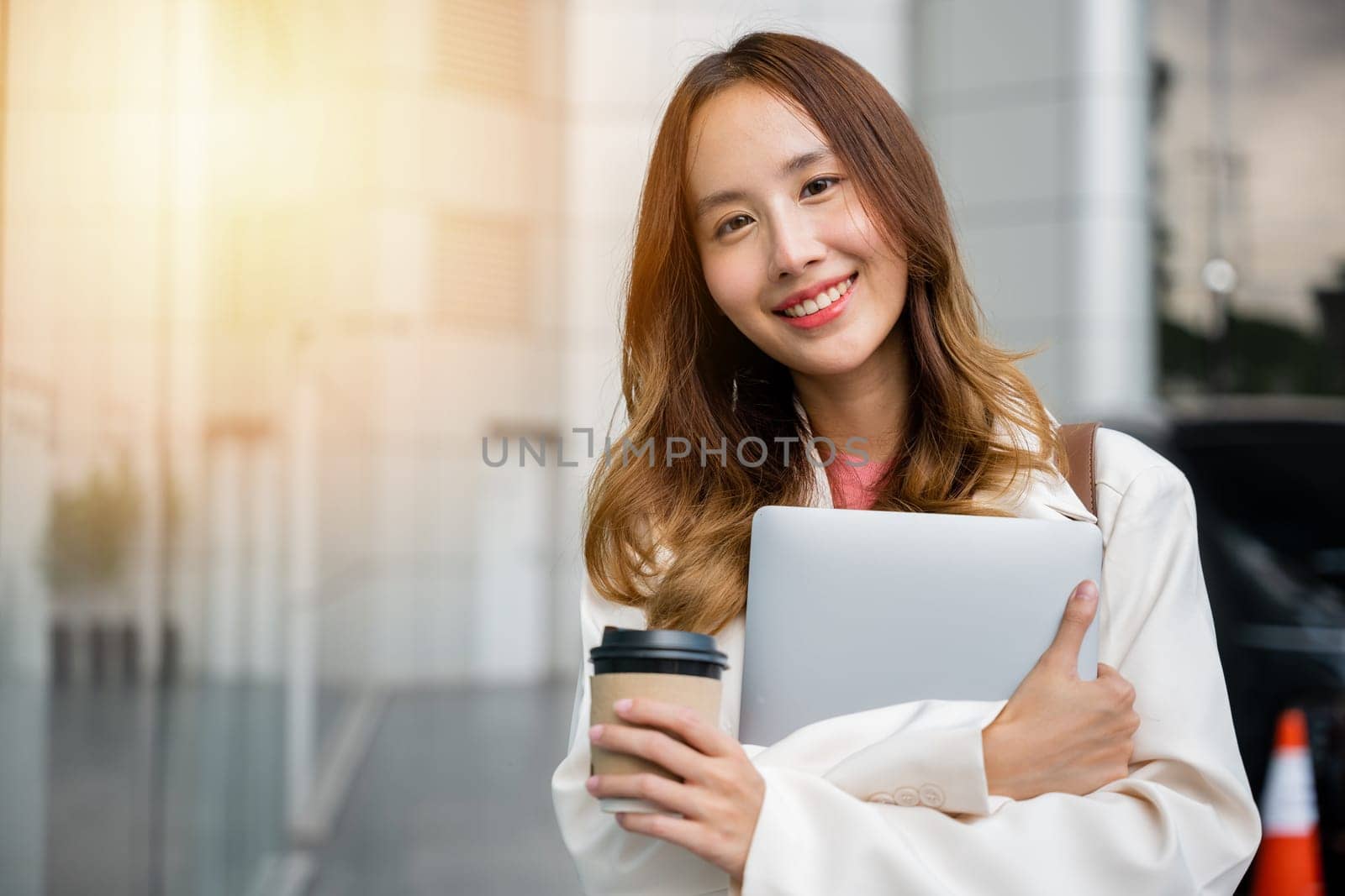 Walking confidently through the city with a cup of takeaway coffee in hand, a brunette woman holds a laptop and smiles at the camera. She embodies the modern-day businesswoman, working and relaxing