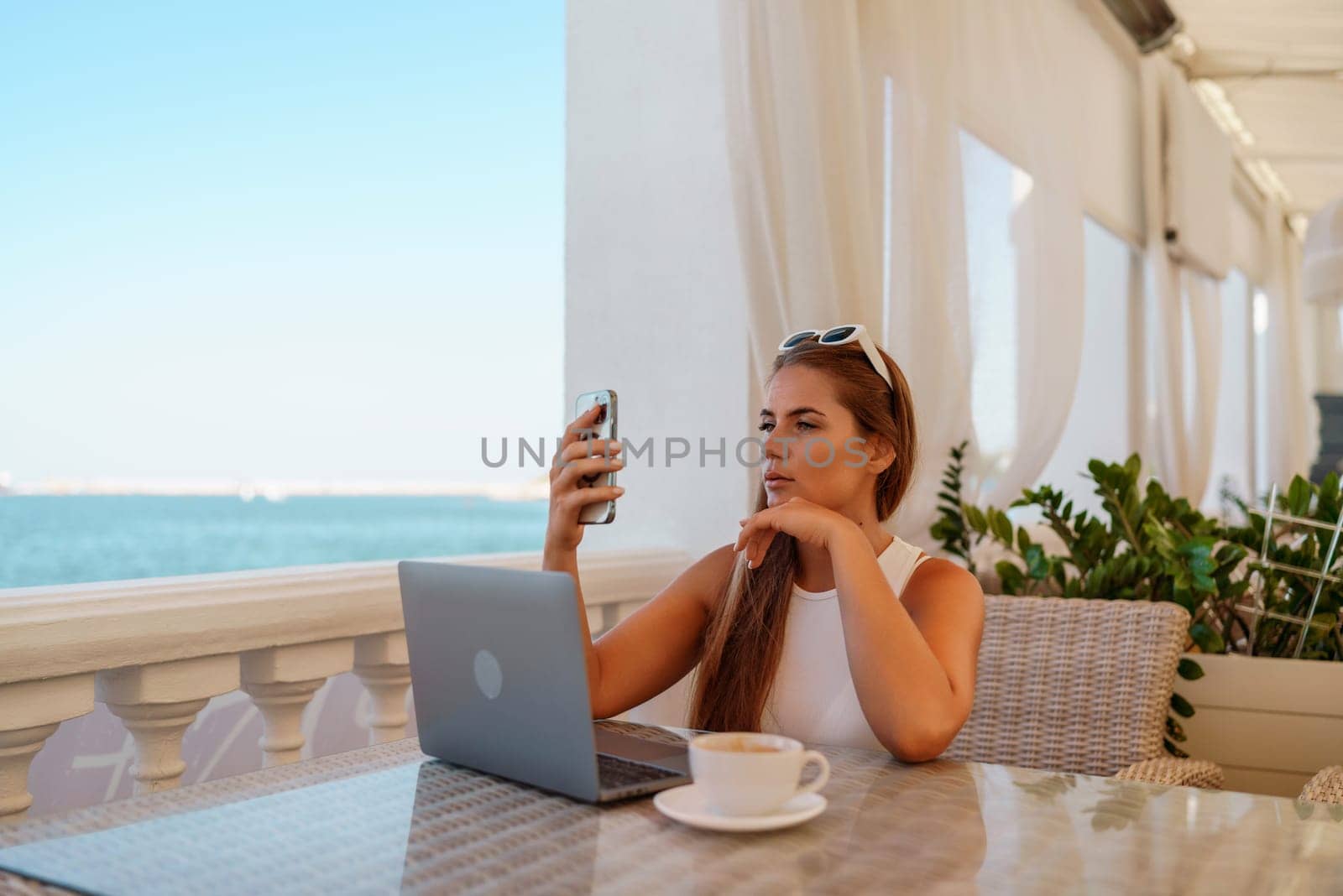 Woman coffee cafe laptop. Coffee break in cafe with sea view. Tranquil long haired woman drinking coffee in plant filled place. Woman sitting at a coffee shop with mobile phone drinking coffee. by Matiunina