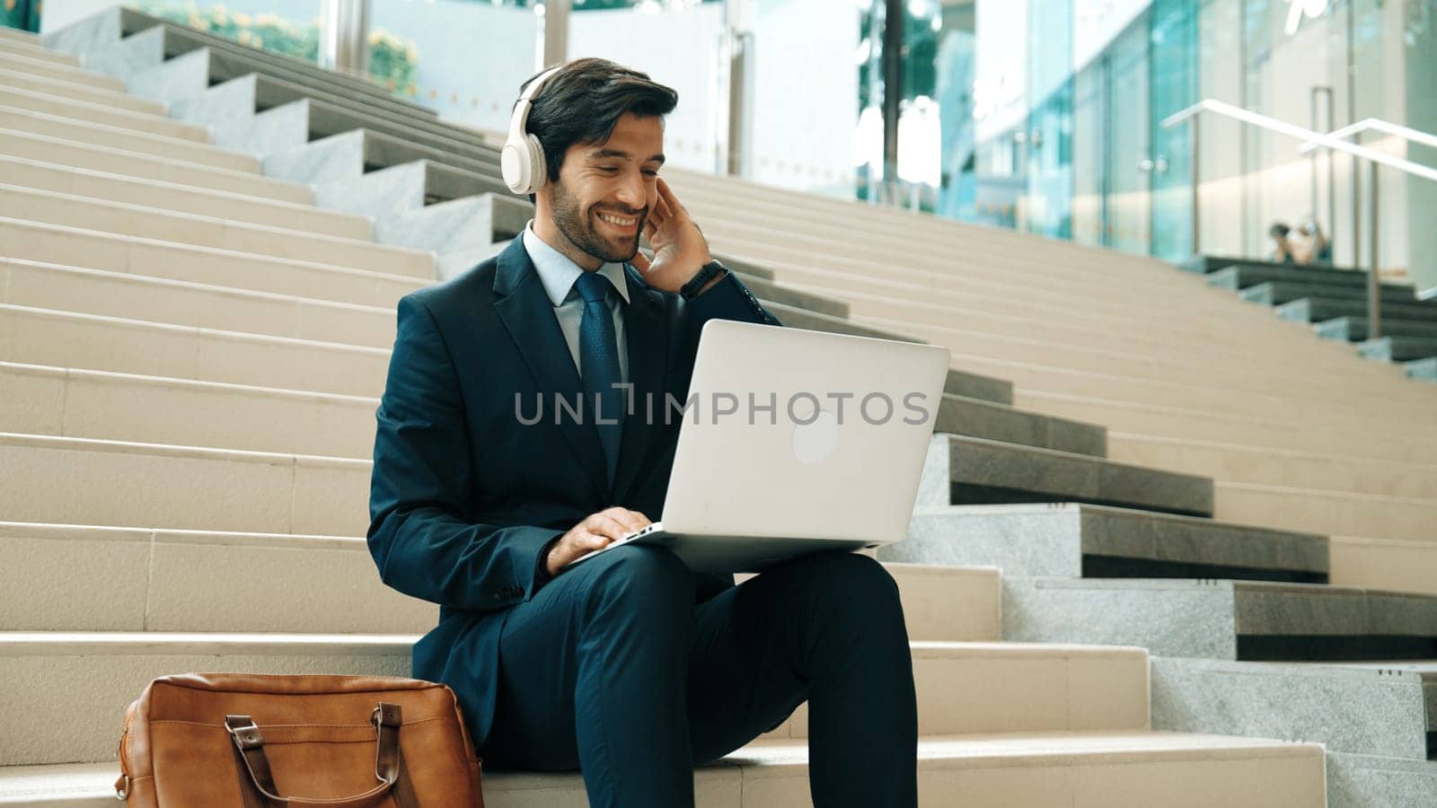 Professional business man sitting at stairs while working on laptop. Skilled project manager listening music from headphone and checking email and discussion about marketing plan. Outdoor. Exultant.