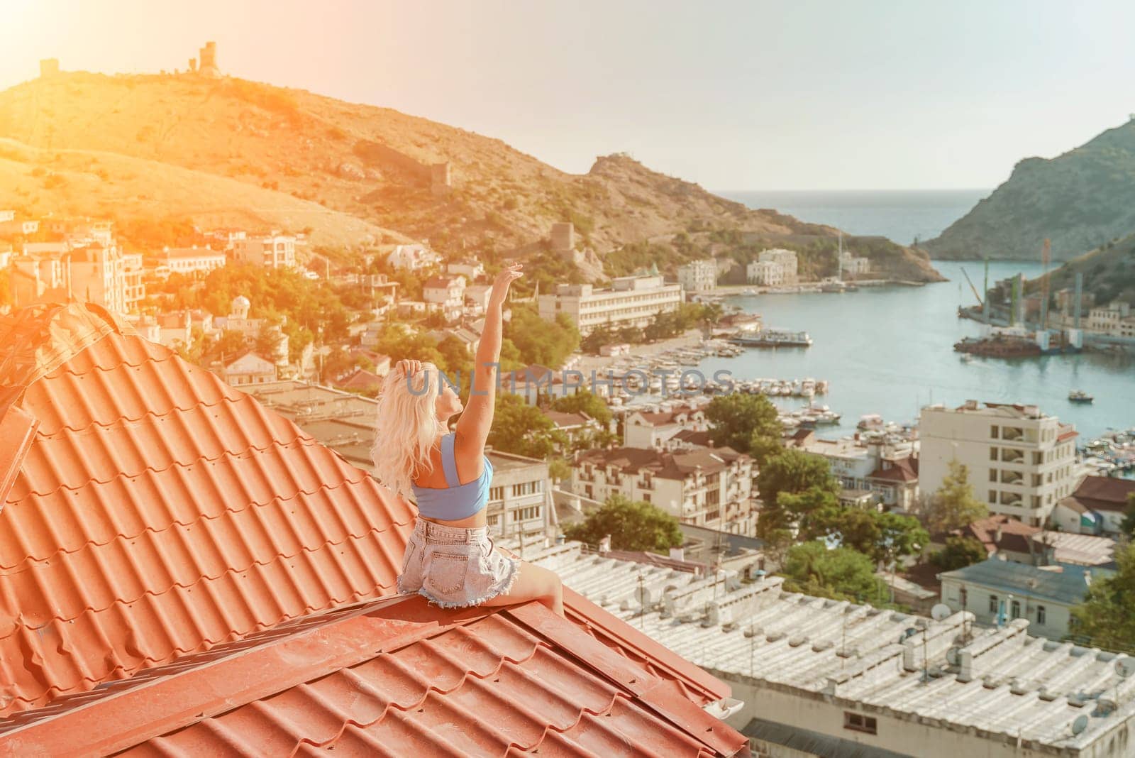 Woman sits on rooftop with outstretched arms, enjoys town view and sea mountains. Peaceful rooftop relaxation. Below her, there is a town with several boats visible in the water by Matiunina