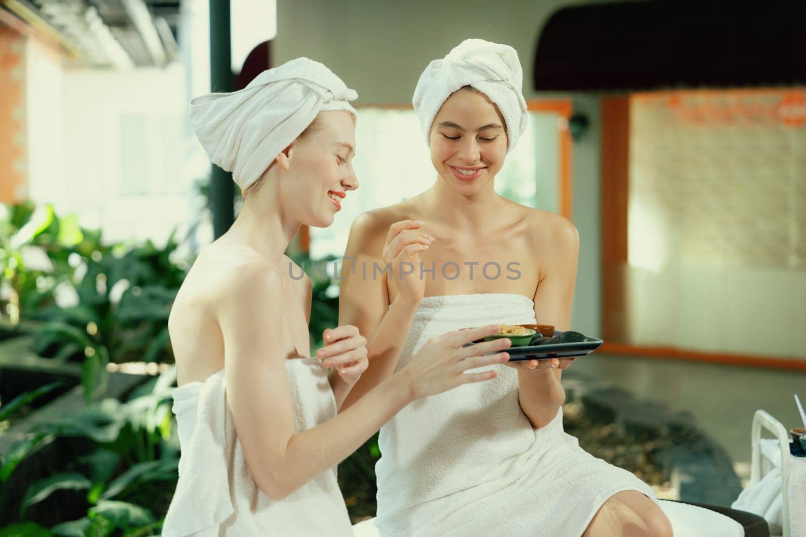 Portrait of two attractive woman in towel giggling during hold the herbal bowl surrounded by natural spa environment. Pretty girls with beautiful skin using herbal scrub at spa salon.Tranquility.