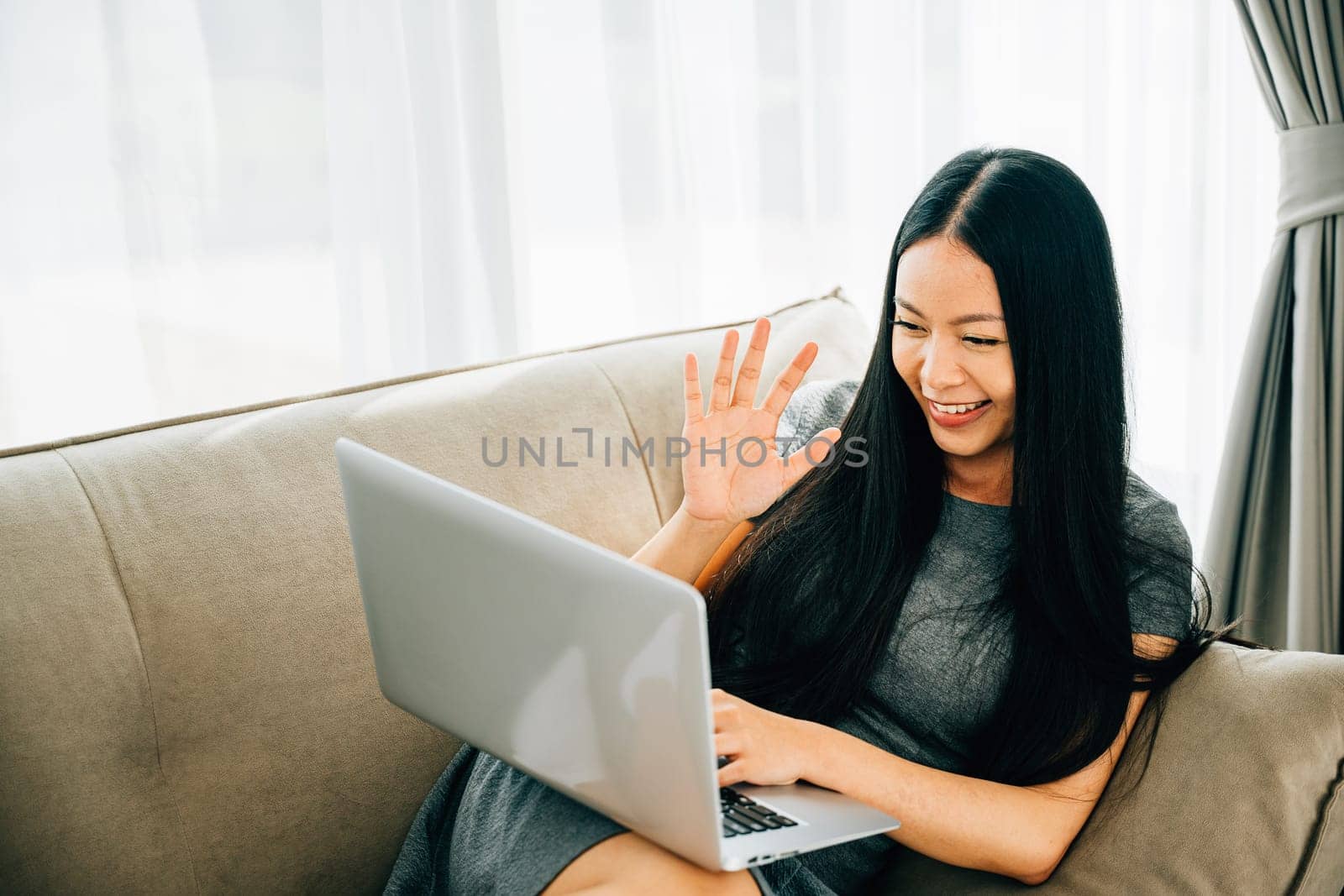 Asian woman on sofa uses laptop for video conferencing greeting colleague by Sorapop