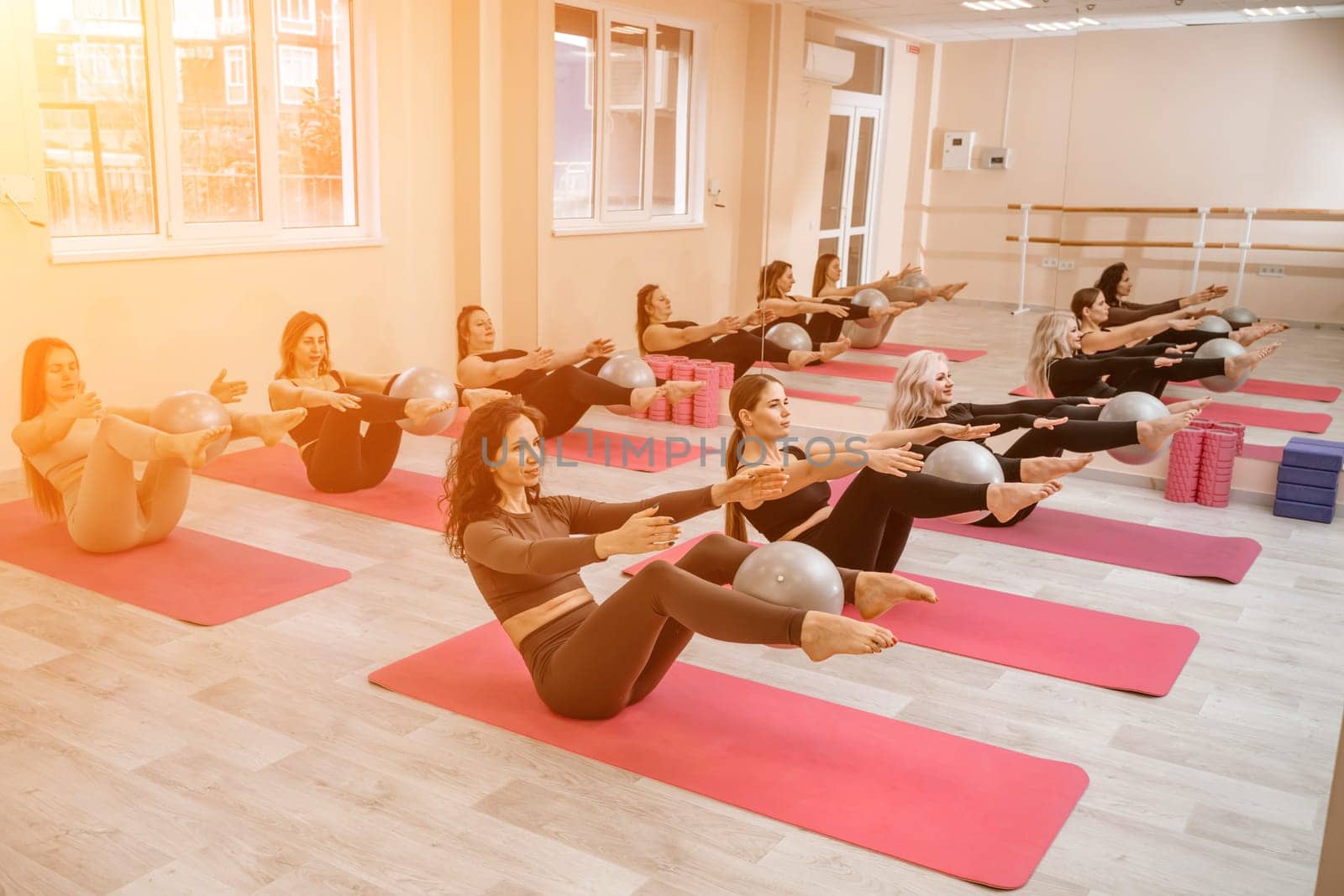 A group of six athletic women doing pilates or yoga on pink mats in front of a window in a beige loft studio interior. Teamwork, good mood and healthy lifestyle concept. by Matiunina