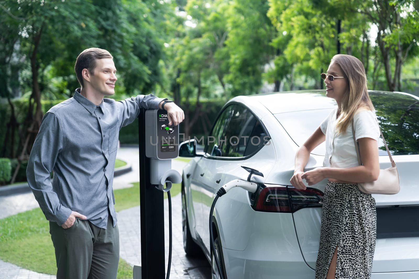 Young couple travel with EV electric car charging in green sustainable city outdoor garden in summer shows urban sustainability lifestyle by green clean rechargeable energy of electric vehicle innards