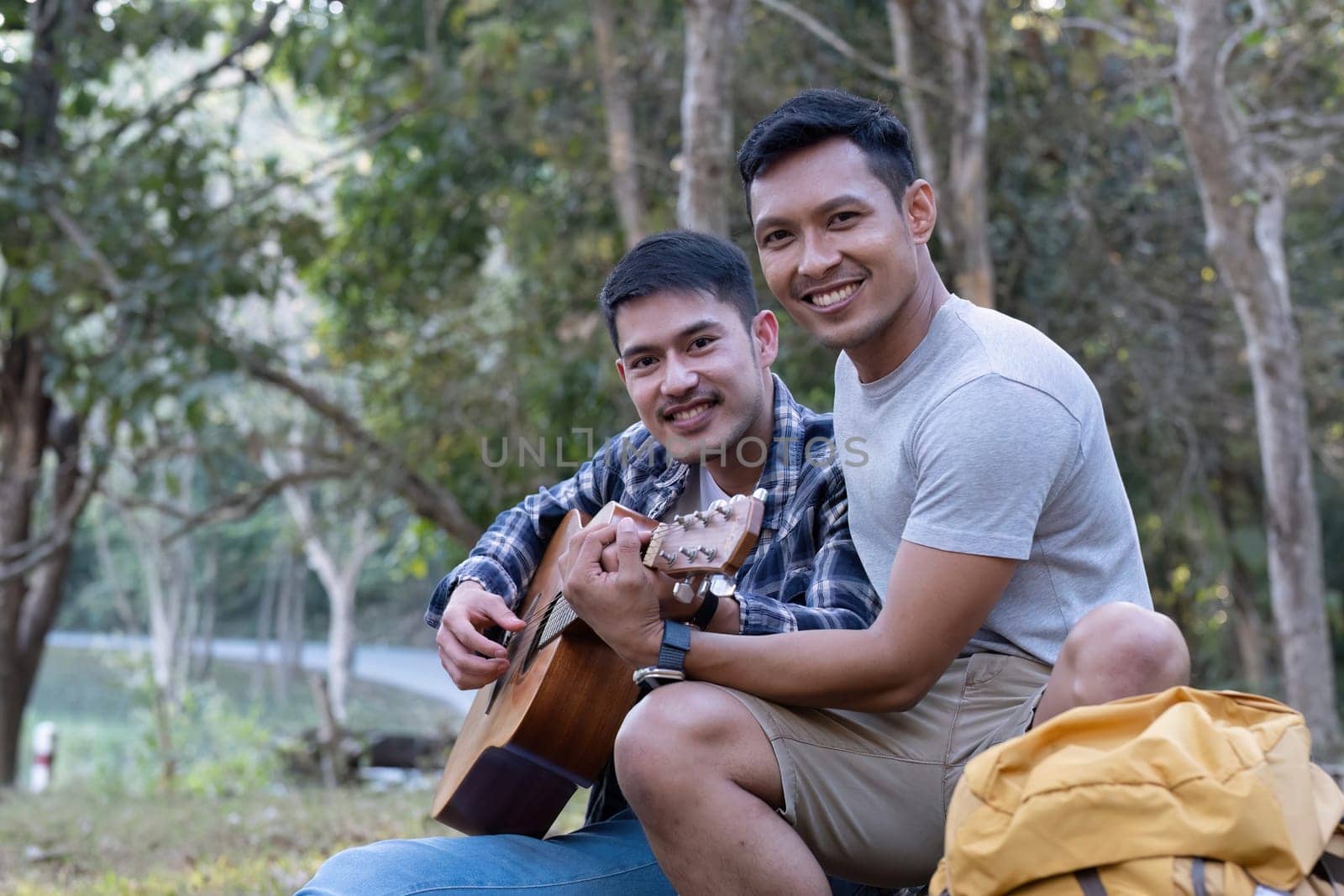 Asian LGBTQ couple enjoying nature, camping with tents in the forest area by the river, playing guitar. by wichayada