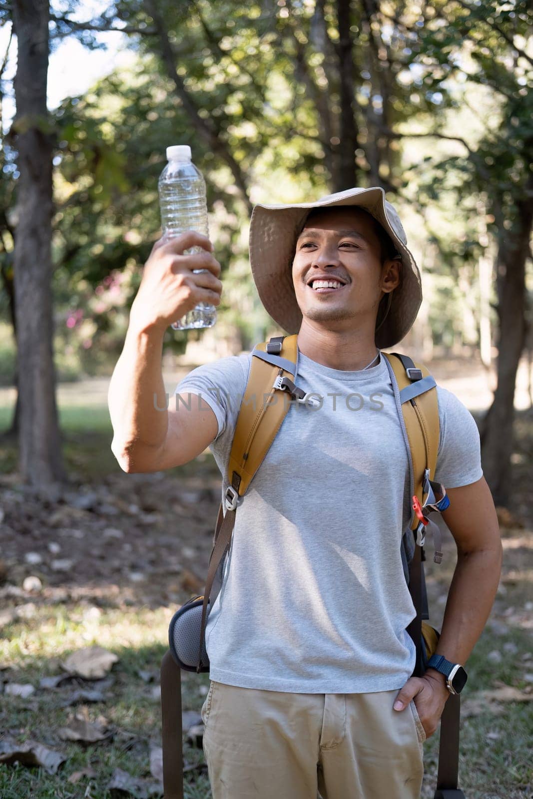 Asian male traveler carrying a large backpack drinks water from a bottle while resting during a hike. by wichayada