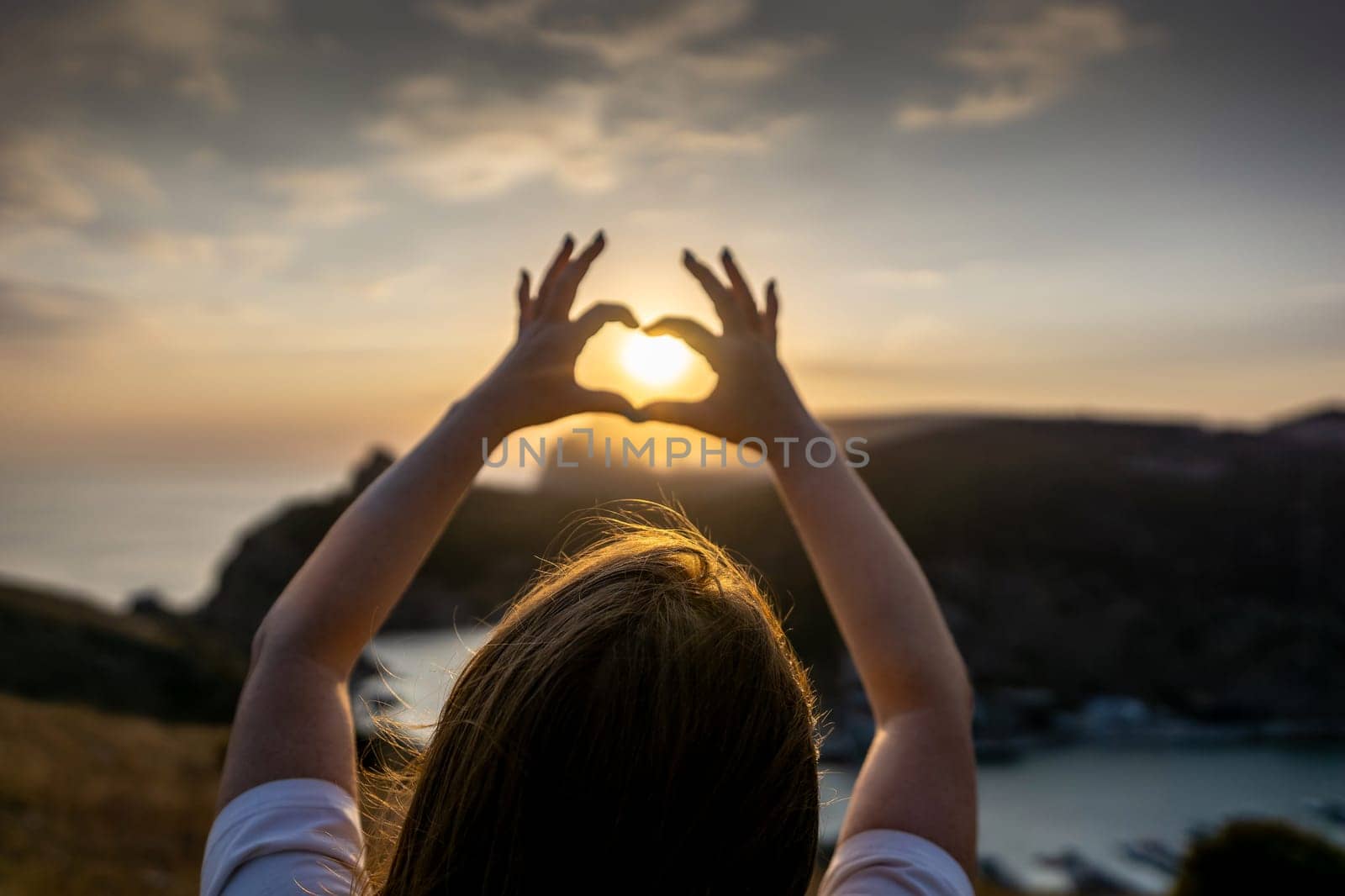 Happy woman on mountain peak, she makes heart shape with hands. Mountain, overlooking sea at sunset. Depicting love shape amidst scenic natural setting