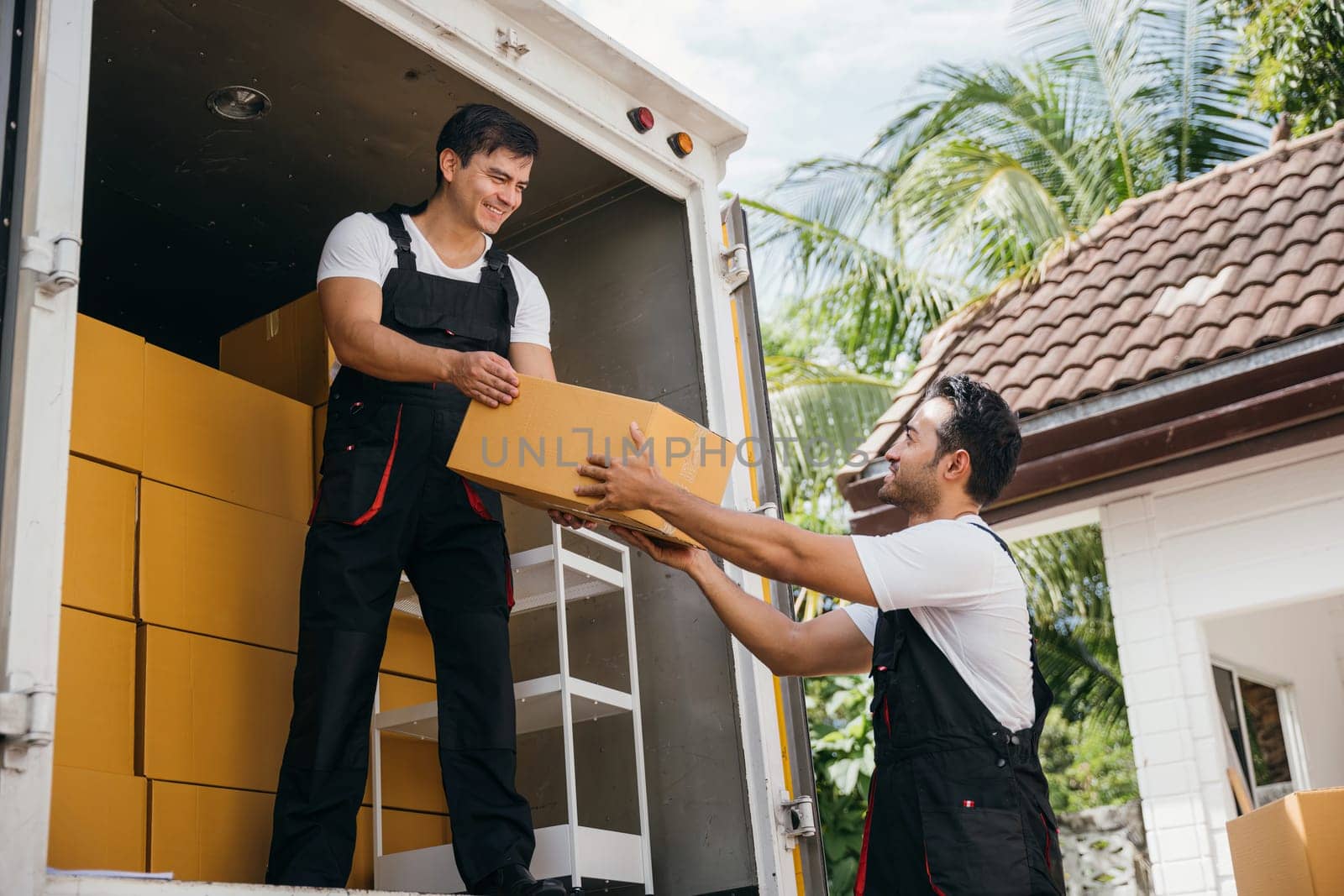 Young delivery men unload boxes from a van. Movers in uniform work together delivering orders. Cooperation in relocation service. Smiling workers carrying boxes. relocation teamwork Moving Day by Sorapop