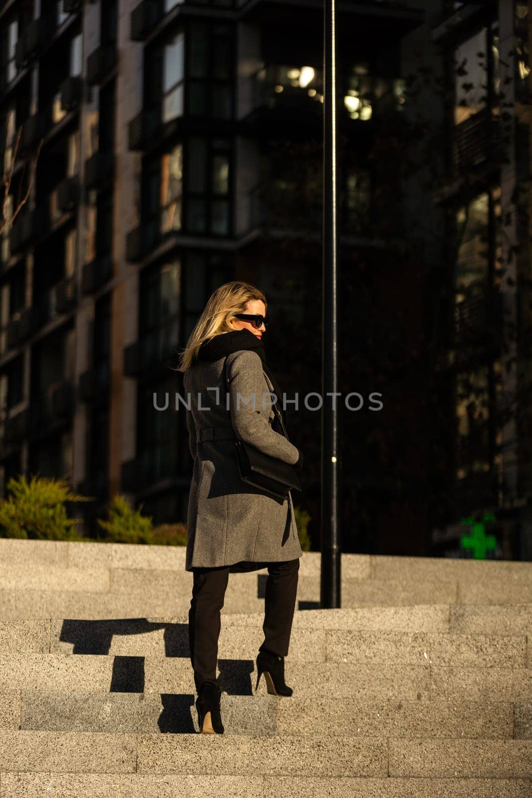 The young lady in black stylish coat with black scarf walking the sun rays at sunset on the background the cold autumn day in the city. High quality photo