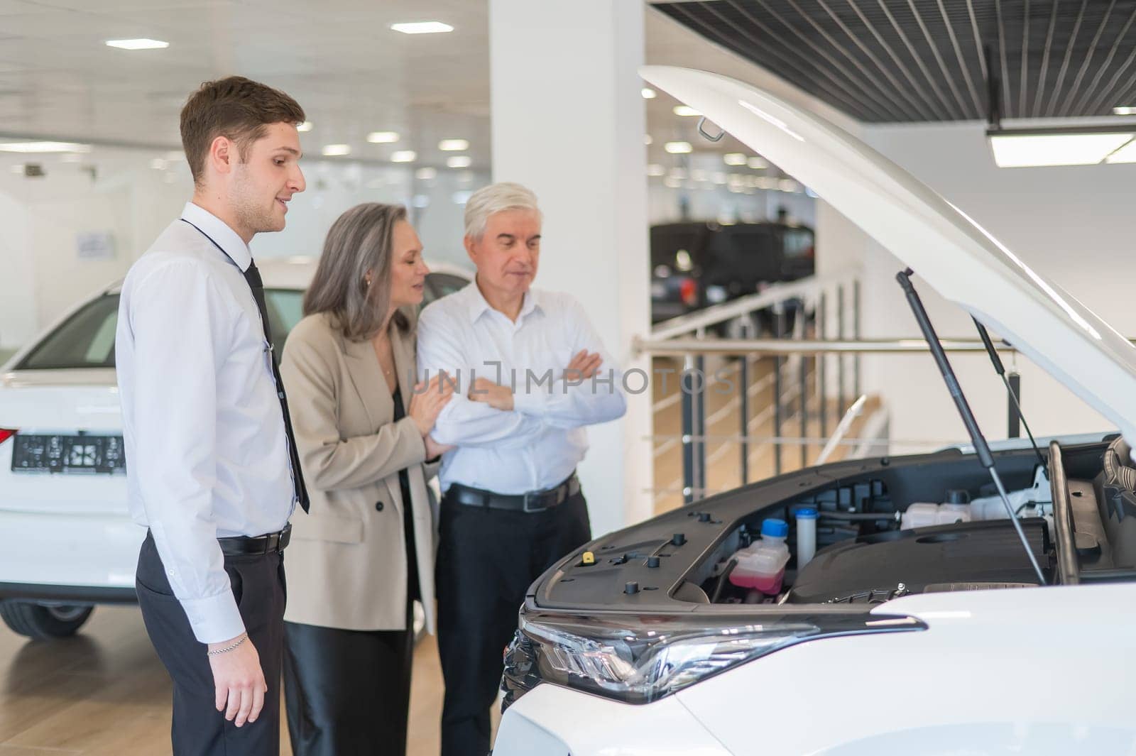 A salesman demonstrates a car with an open hood to an elderly Caucasian couple in a car dealership. by mrwed54
