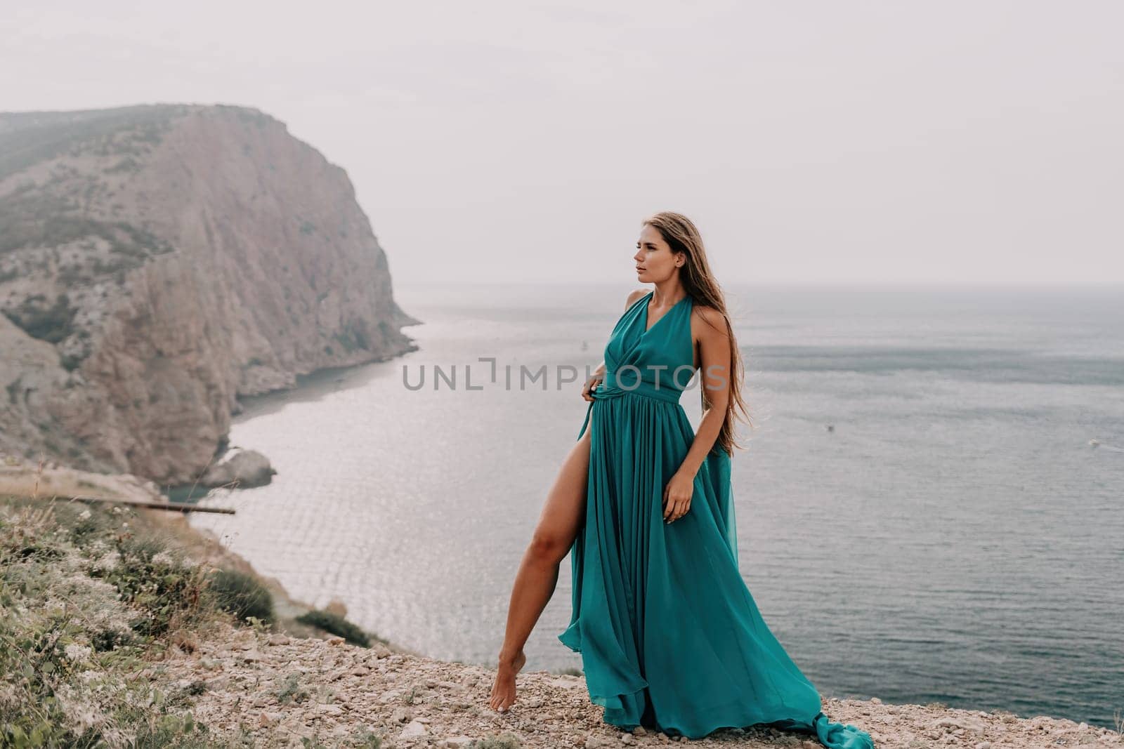 Woman travel portrait. Happy woman with long hair looking at camera and smiling. Close up portrait cute woman in a mint long dress posing on a volcanic rock high above the sea by panophotograph
