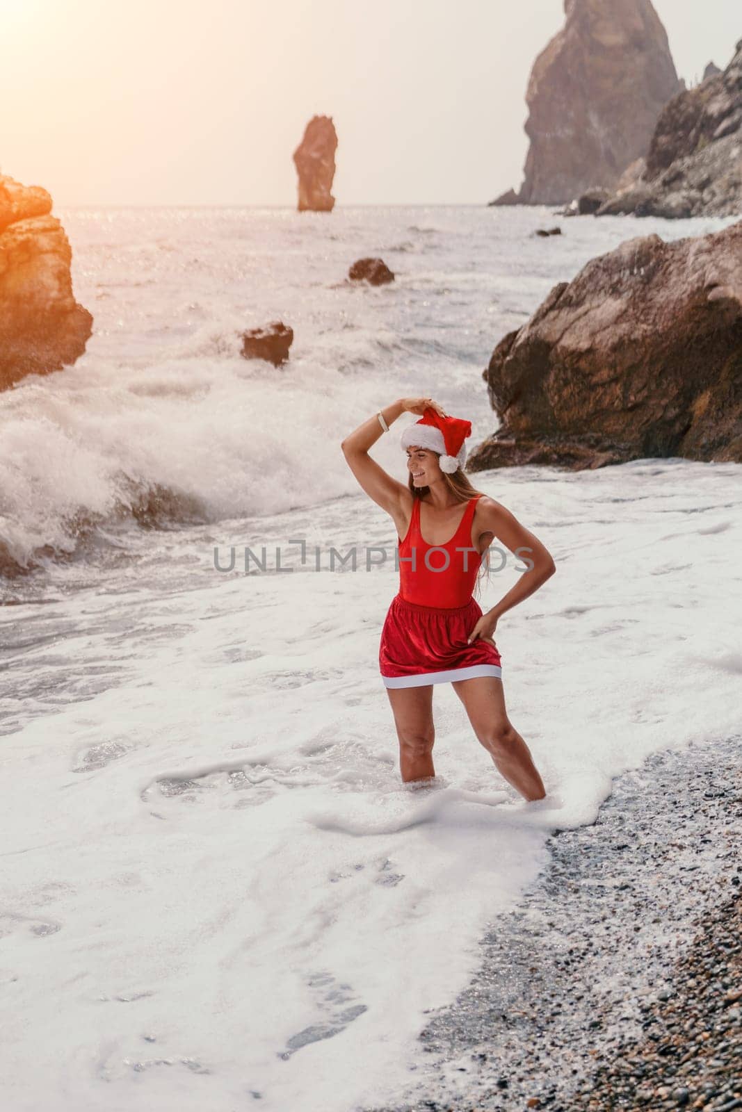 Woman summer travel sea. Happy tourist in red bikini and Santas hat enjoy taking picture outdoors for memories. Woman traveler posing on the beach surrounded by volcanic mountains, sharing travel joy by panophotograph