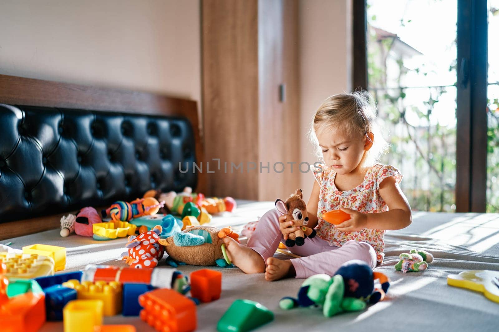 Little girl plays with a plush deer on the bed among colorful soft toys. High quality photo