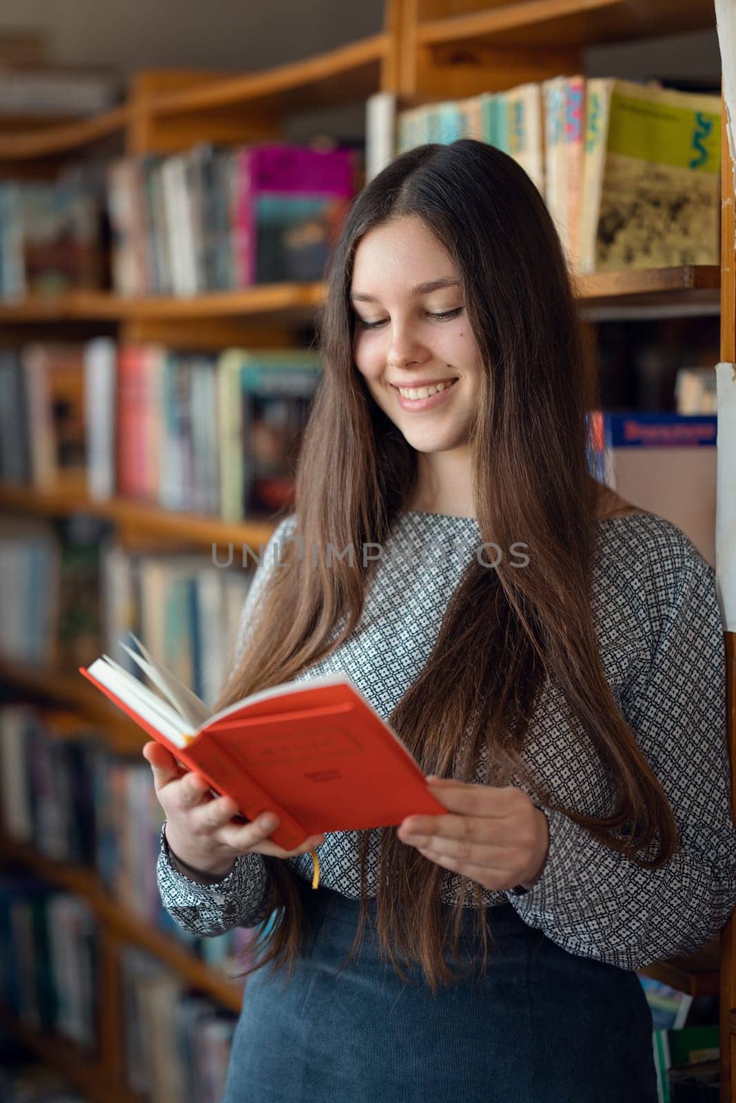 Attractive female brunette student standing with a book in her hands near the rows of bookshelves in library by VitaliiPetrushenko