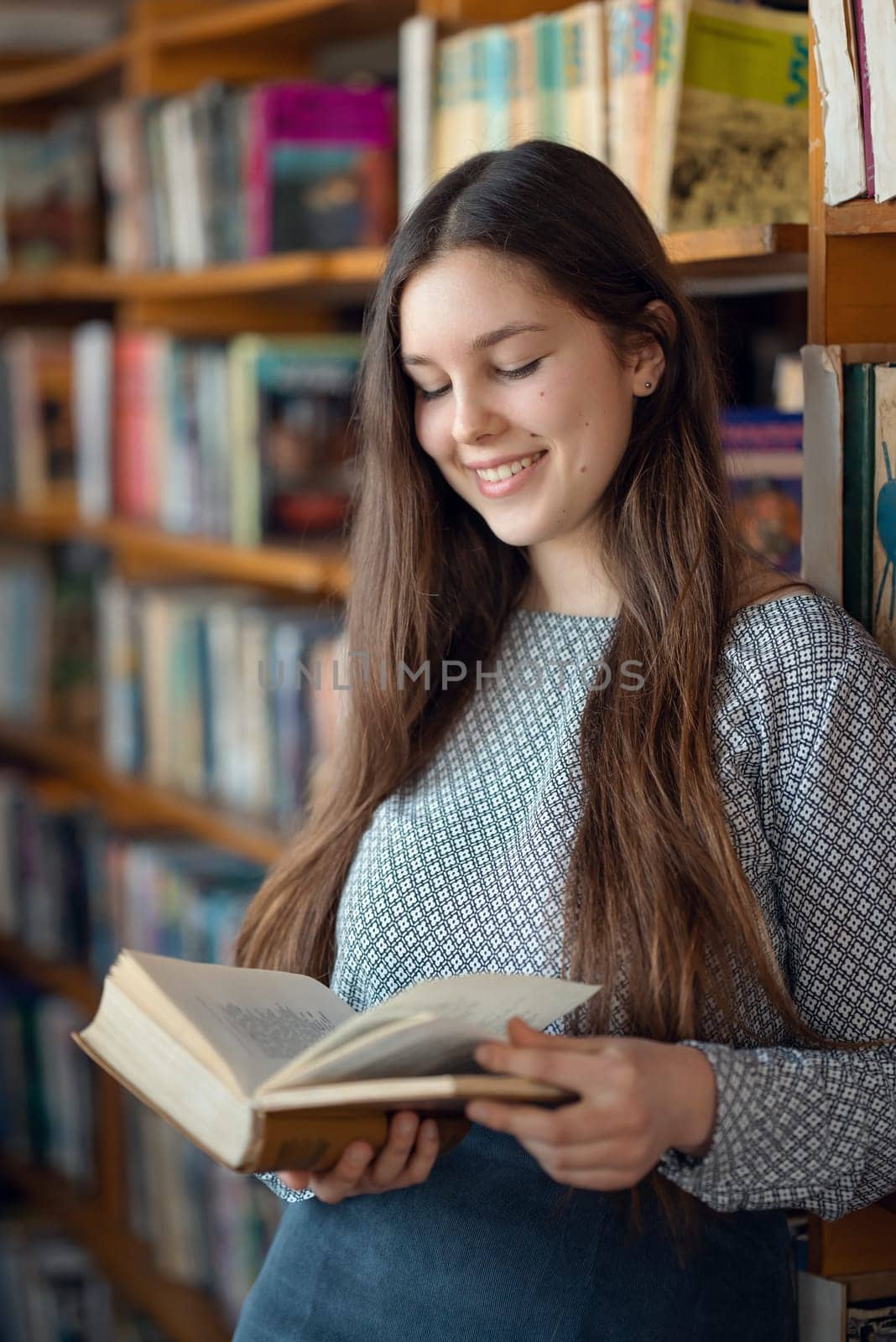 Cheerful relaxed girl standing with book in her hands, library shelves on the background
