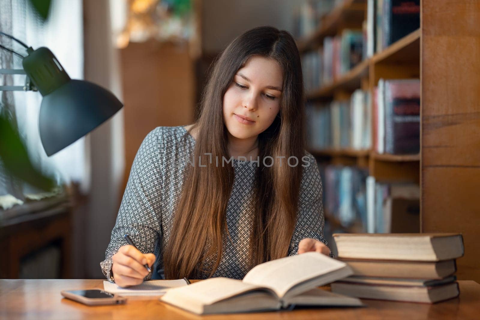 Concentrated young female student sitting at the table, reading and writing something for her studying by VitaliiPetrushenko