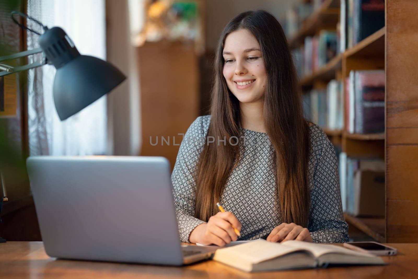 Cute young student girl sitting at a table with laptop in front of her, writing something in the notebook by VitaliiPetrushenko