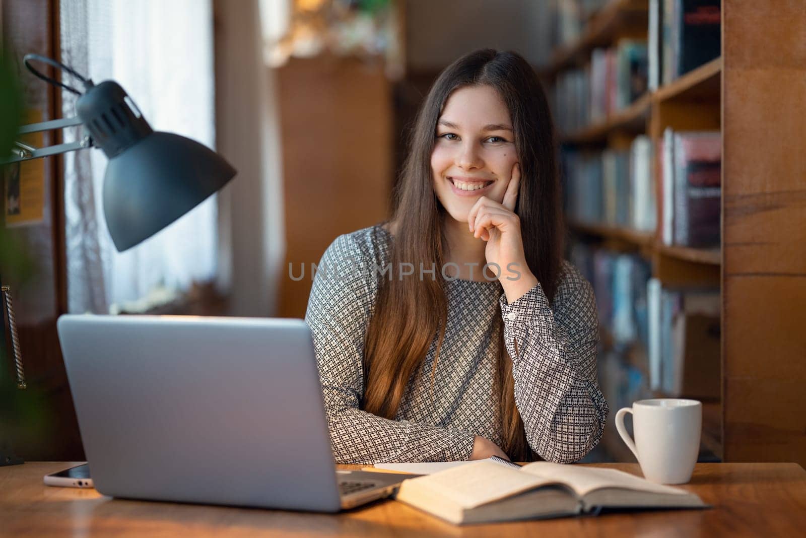 Cheerful female student sitting at a table in library with laptop, book and cup of tea in front of her by VitaliiPetrushenko