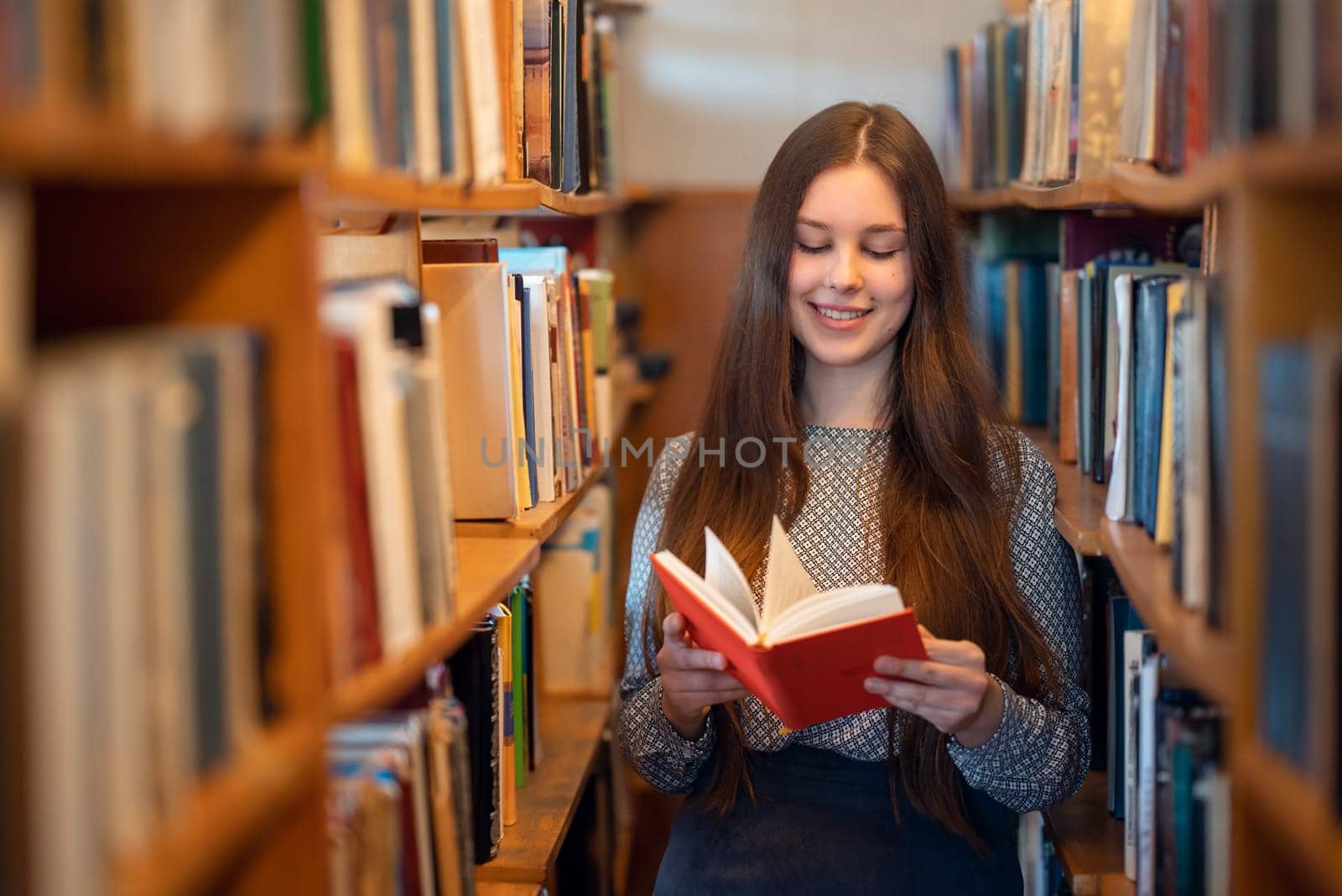 Girl reading a book with red cover in the library by VitaliiPetrushenko
