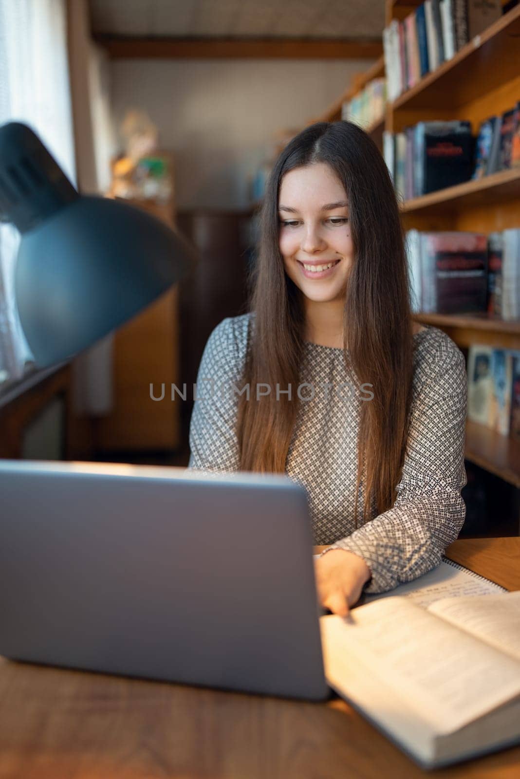 Vertical shot of an intelligent young brunette girl studying in a library, sitting at a table with laptop in front of her by VitaliiPetrushenko