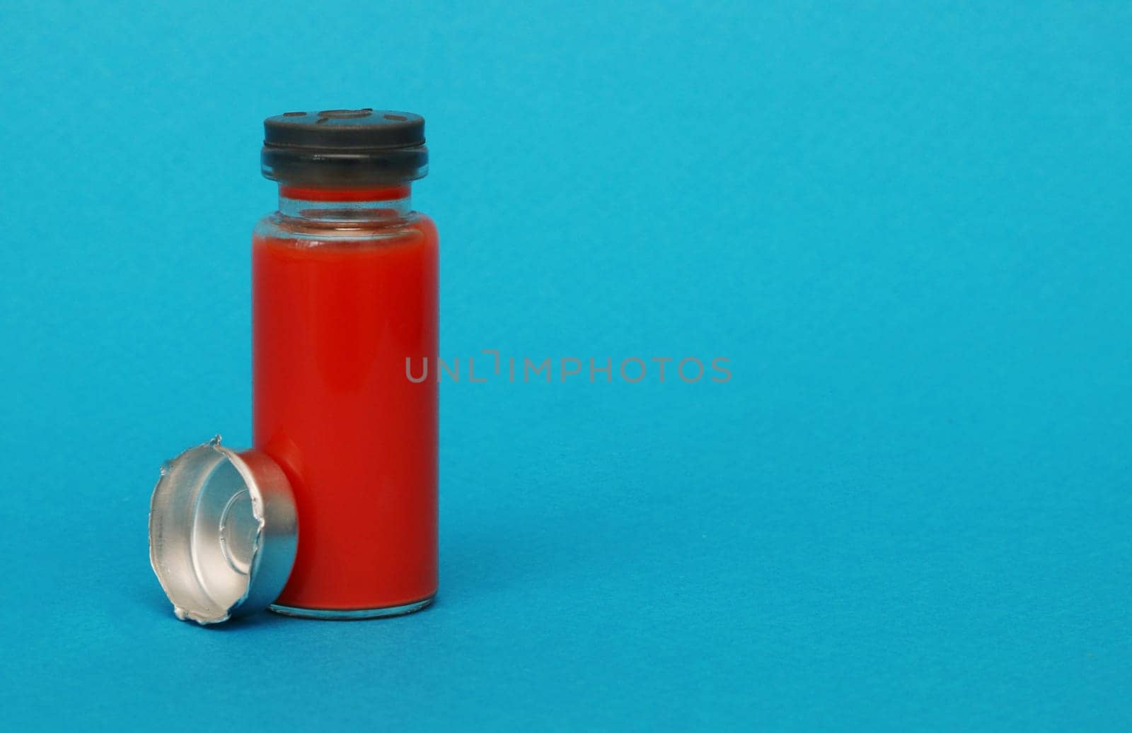 Glass medical bottle with a metal cap filled with blood. Bottle with red liquid on a blue paper background.