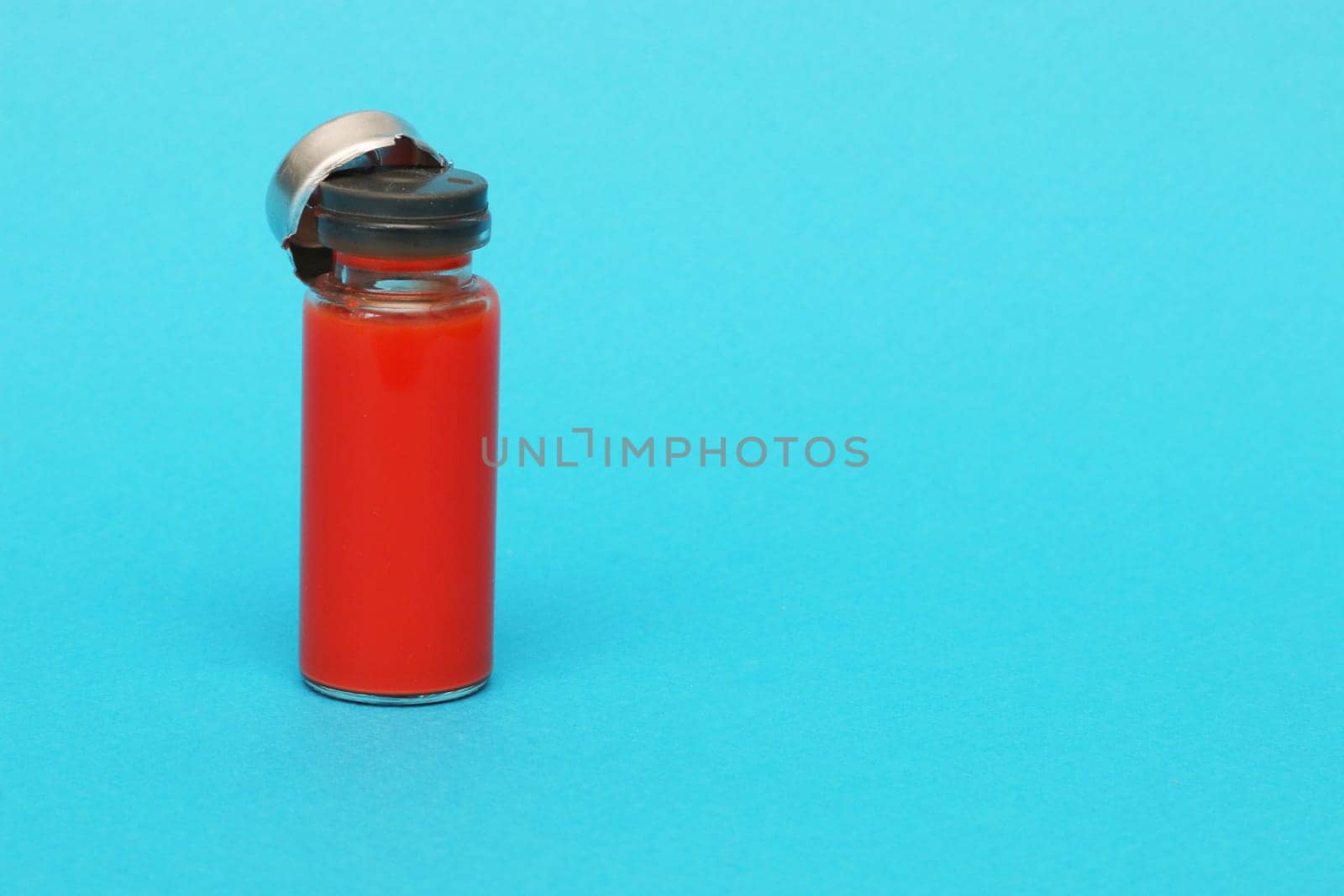 Glass medical bottle with a metal cap filled with blood. Glass bottle with red liquid on a blue paper background.