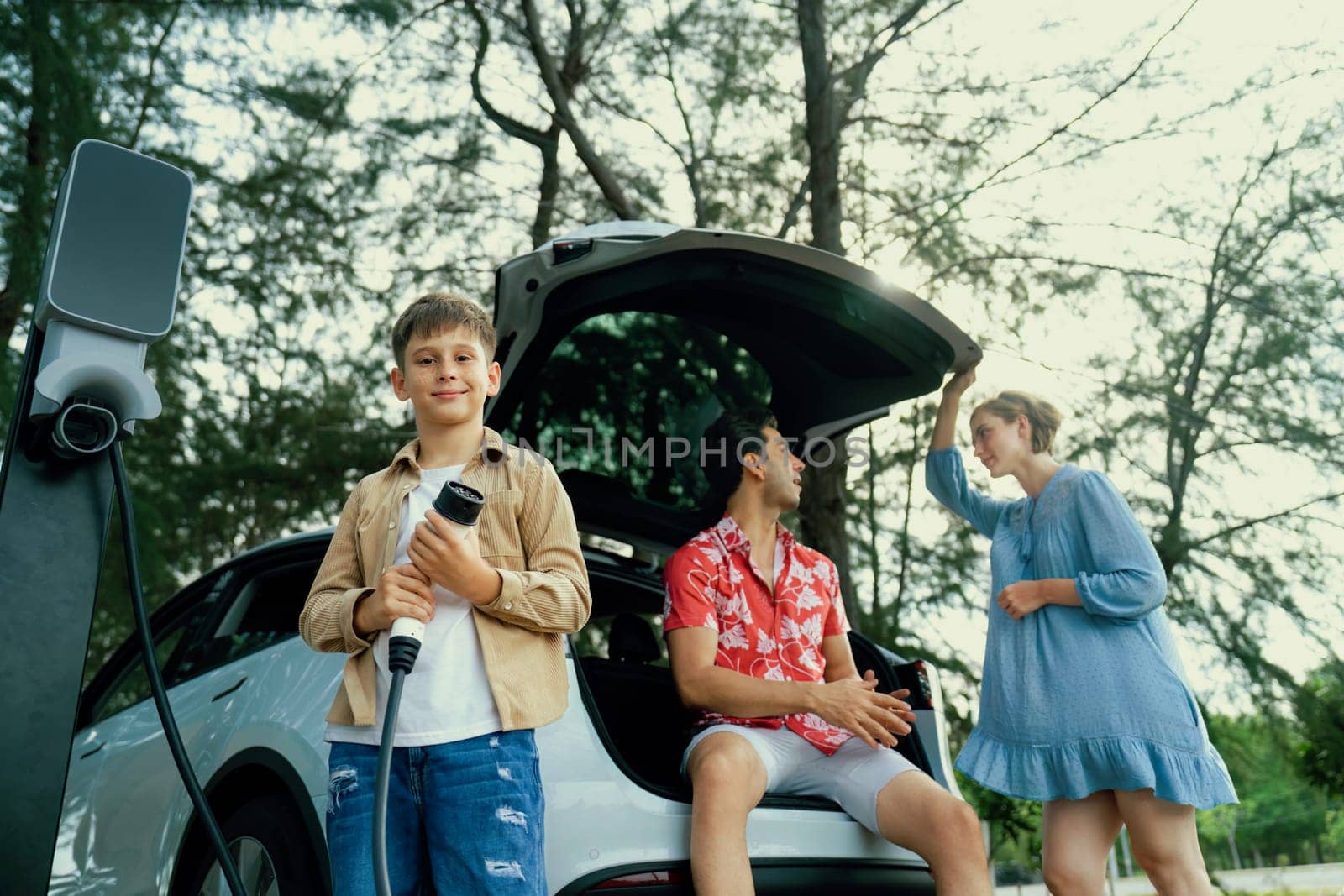 Little boy holding EV charger and point at camera with his family. Perpetual by biancoblue