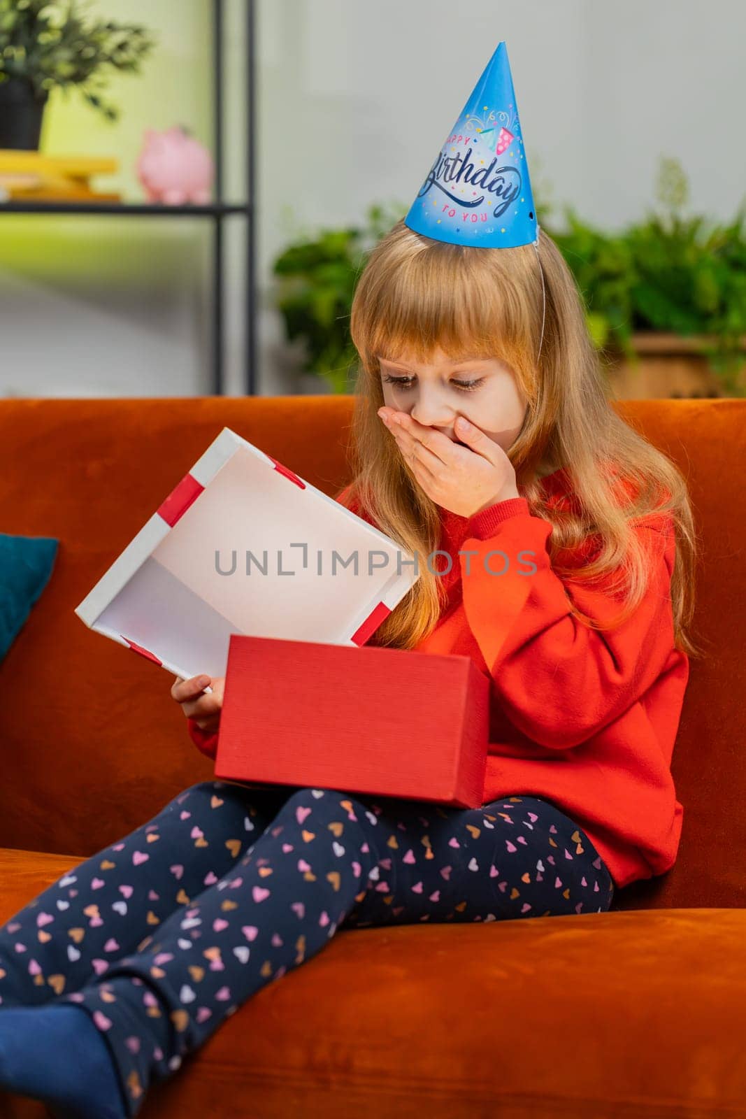 Happy preteen child girl wears festive birthday cap hat hold gift box with ribbon congratulating. Teenager kid celebrating party event, opening delivery greeting present at home room on sofa. Vertical