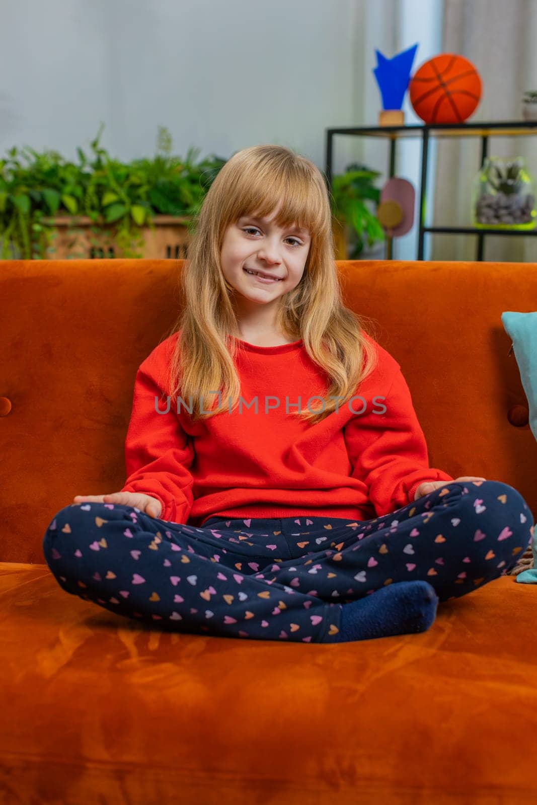 Close-up portrait of happy smiling cheerful Caucasian preteen school girl 8 years old. Young lovely blonde little child kid looking at camera at home play living room sitting on orange couch. Vertical