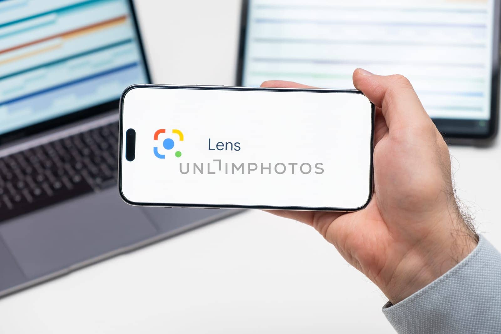 Lens logo of app on the screen of mobile phone held by man in front of the laptop and tablet by vladimka