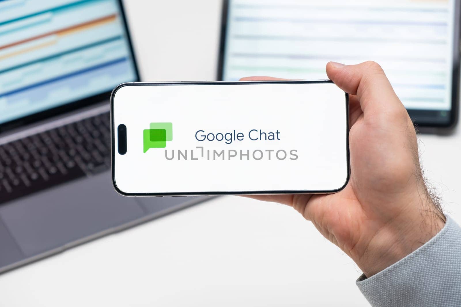 Google Chat logo of app on the screen of mobile phone held by man in front of the laptop and tablet by vladimka