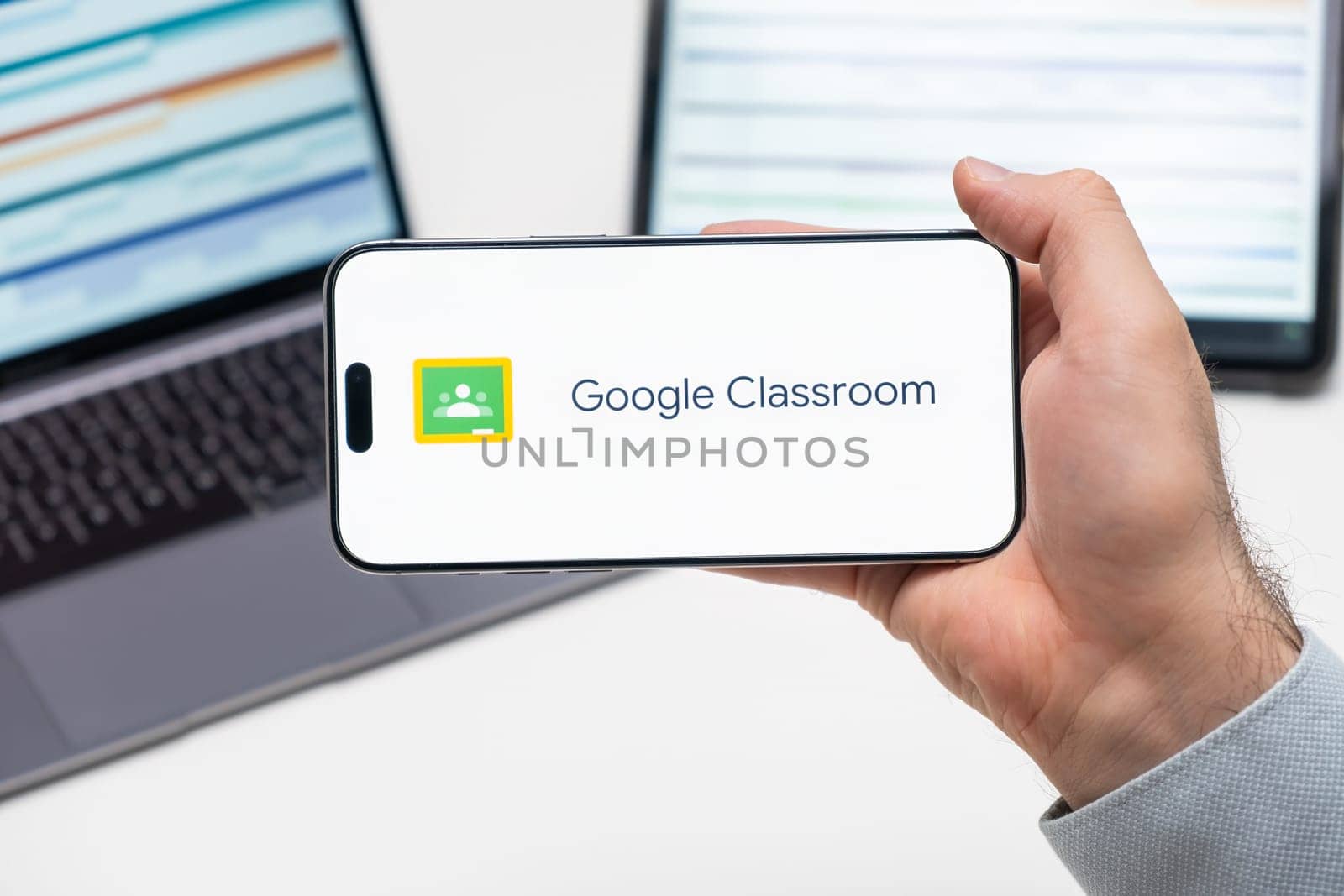 Google Classroom application logo on the screen of smart phone in mans hand, laptop and tablet on the table by vladimka