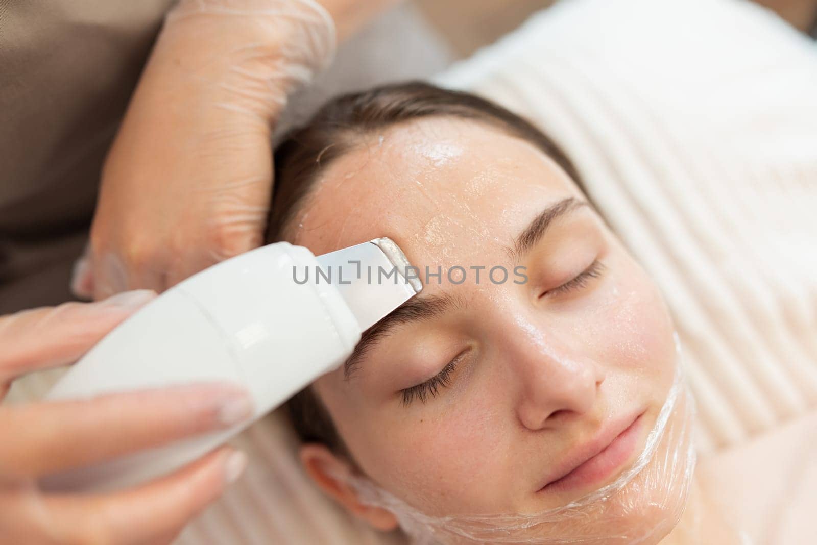 Close up of professional ultrasound cleaning appliance in hands of a cosmetologist cleaning face of a young female client by VitaliiPetrushenko