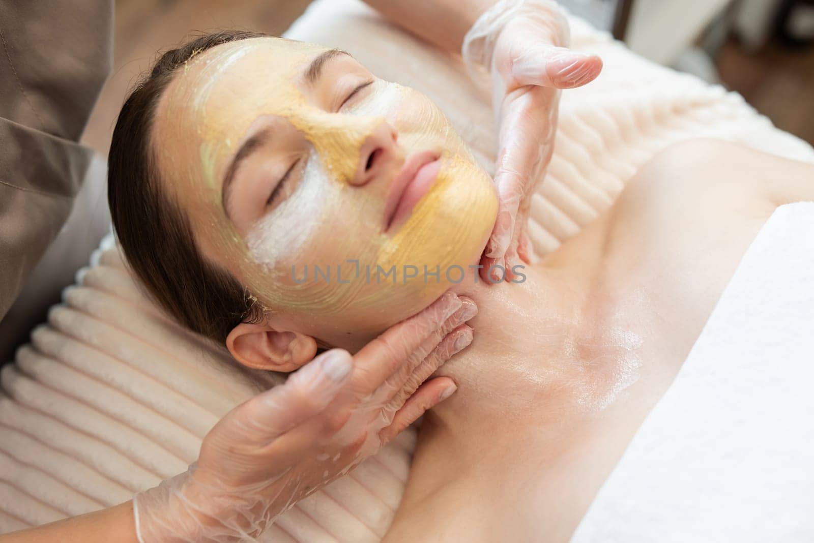 Young girl enjoying spa procedures with moisturizing, cleansing, brightening masks, while cosmetology specialist applying them to her face and neck by VitaliiPetrushenko