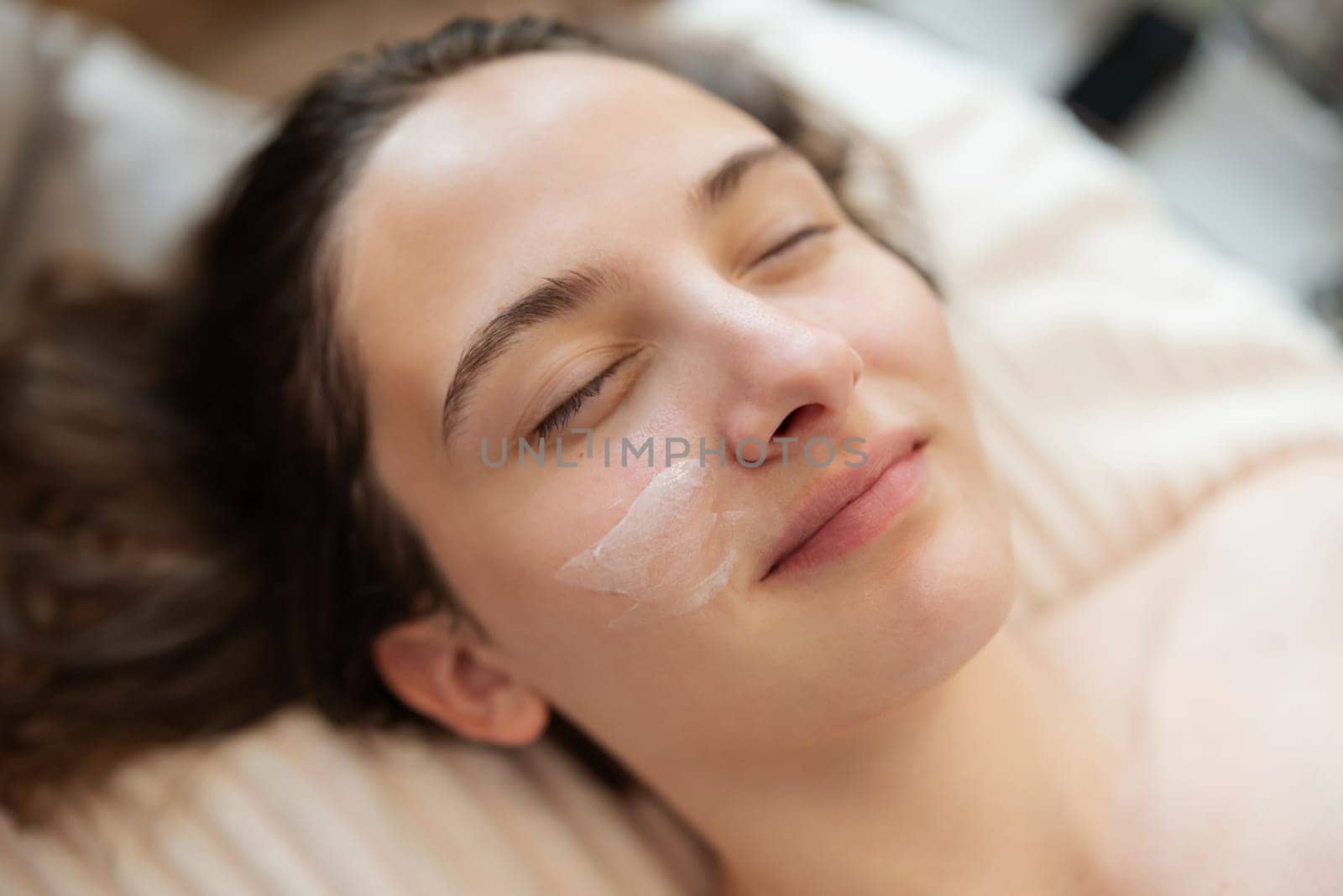 Close up of girl with cream on her cheek laying in bed in cosmetological clinic by VitaliiPetrushenko
