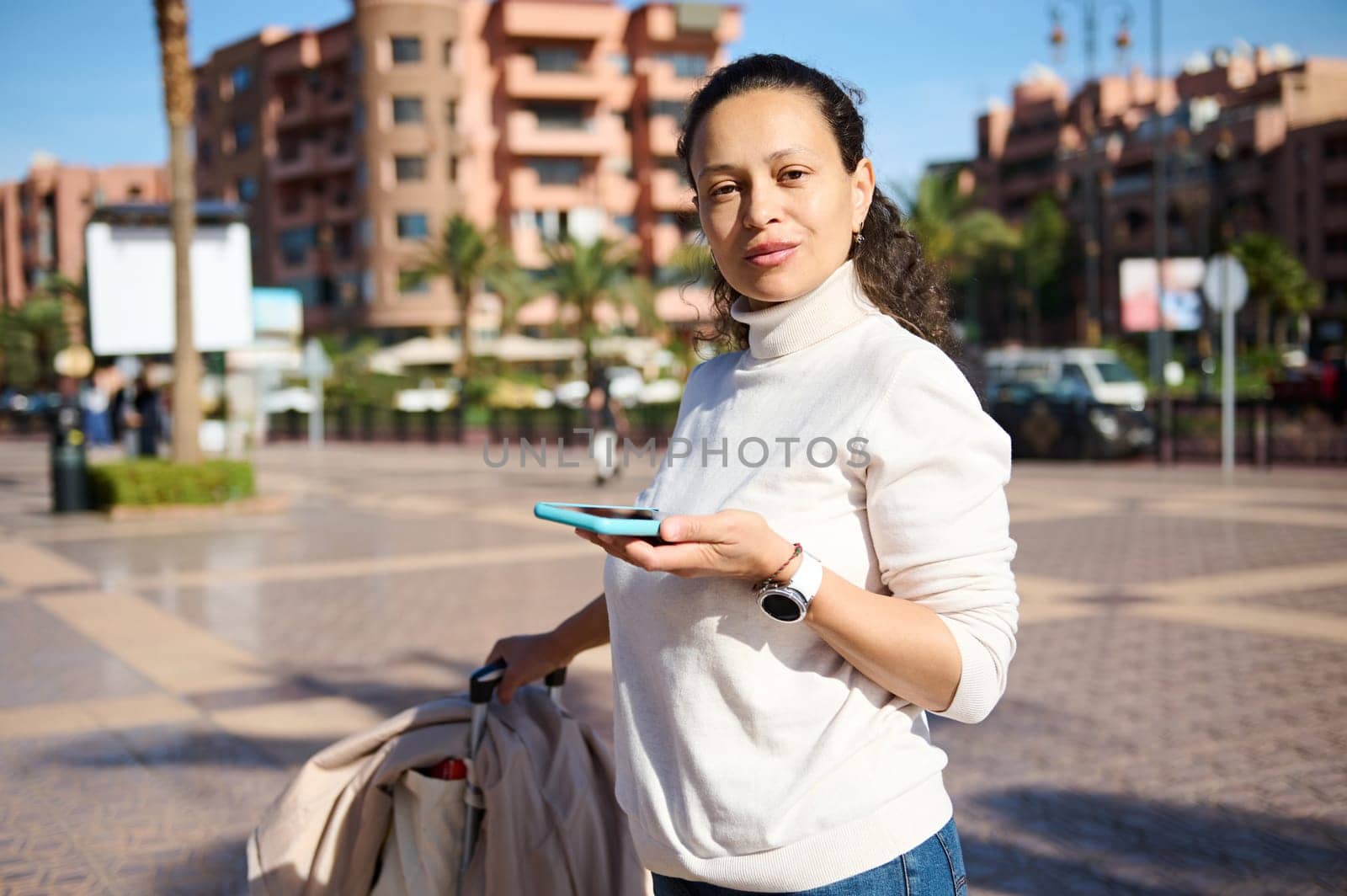 Pretty woman traveler with suitcase, in casual wear using smart mobile phone outdoor, confidently looking at the camera by artgf