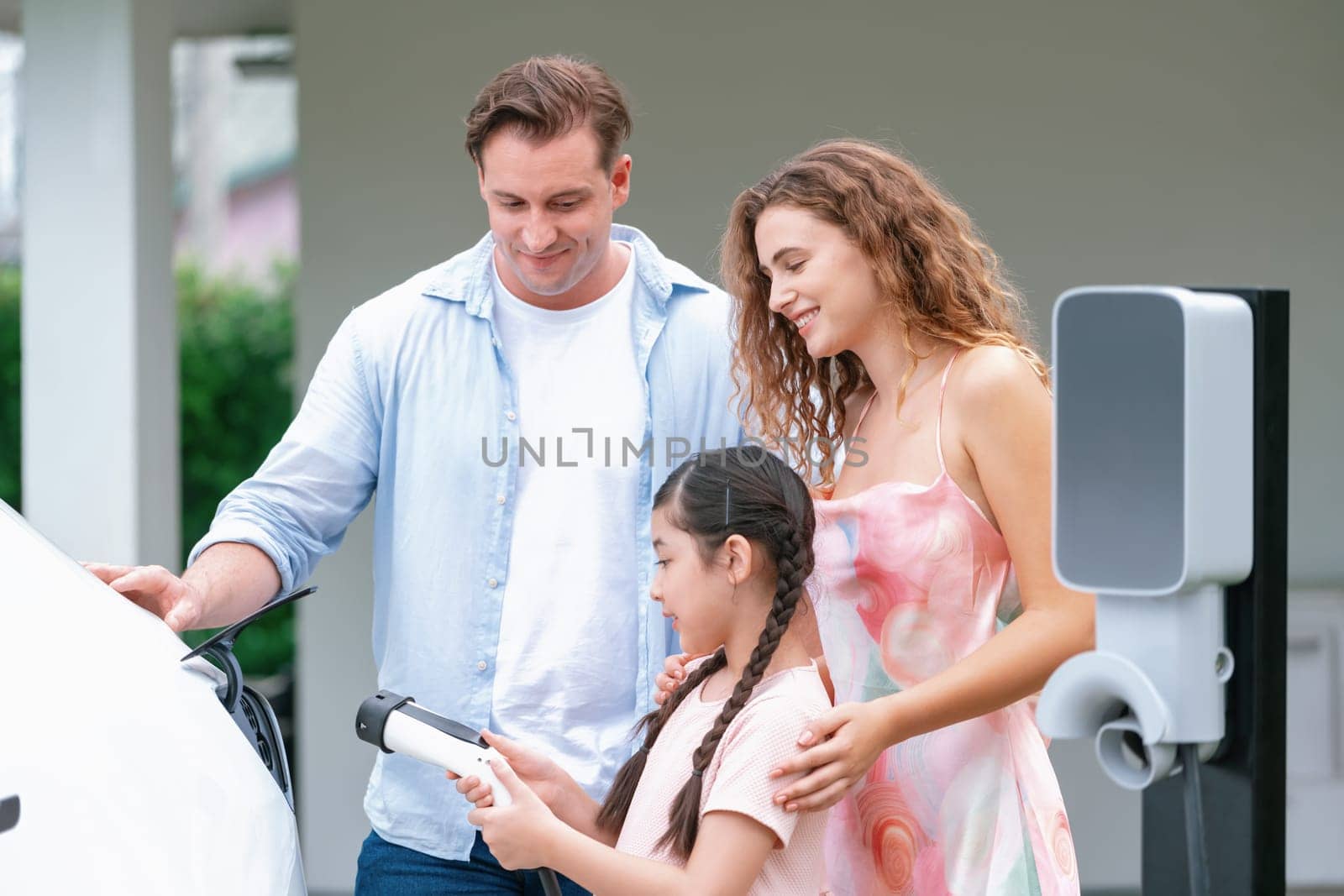 Happy little young girl learn about eco-friendly and energy sustainability as she help her family recharge electric vehicle from home EV charging station. EV car and modern family concept. Synchronos