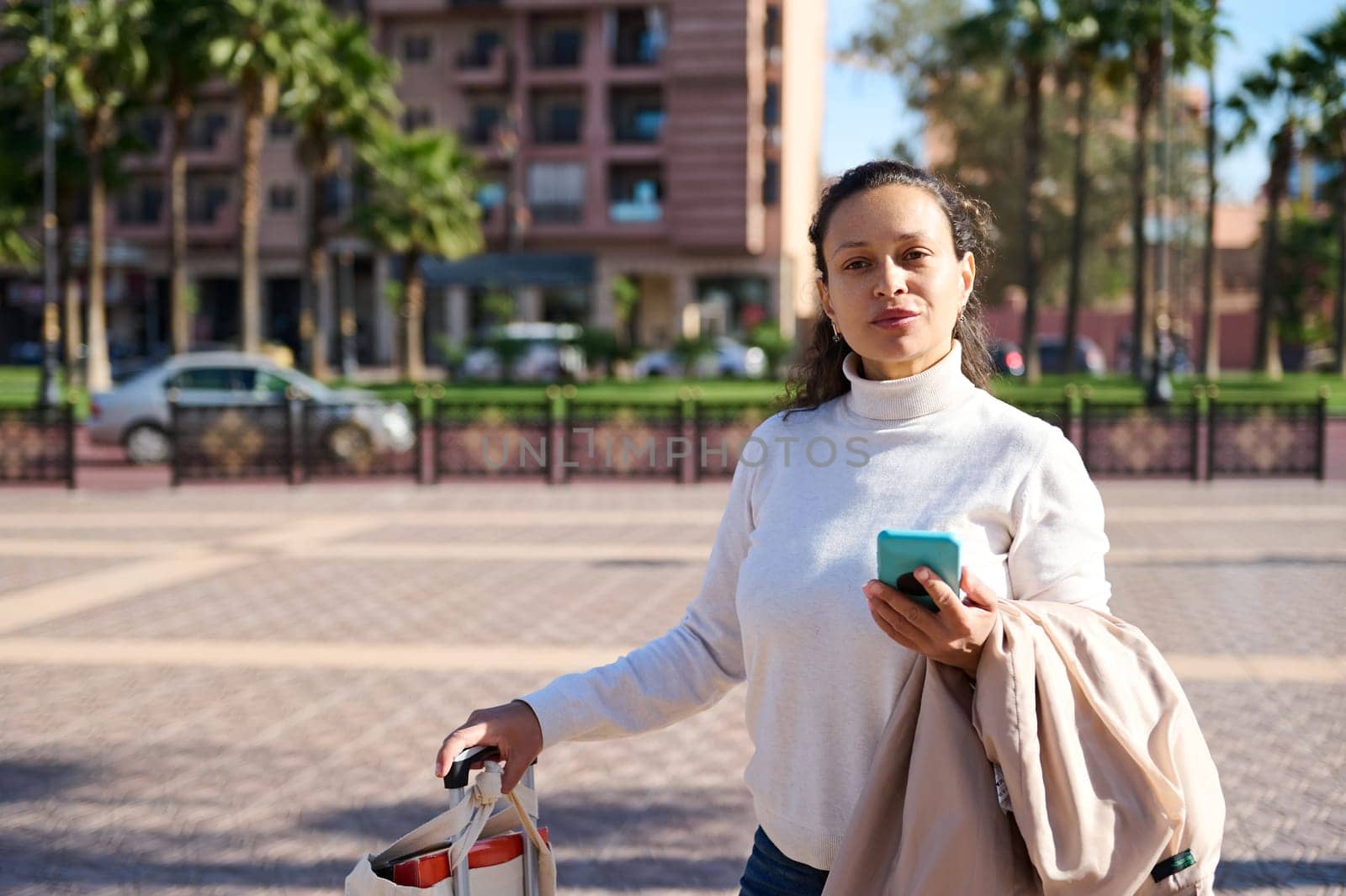 Beautiful African American woman, tourist carrying a suitcase and holding mobile phone while standing on the city square, smiling cutely looking at the camera. People. Lifestyle. Technology