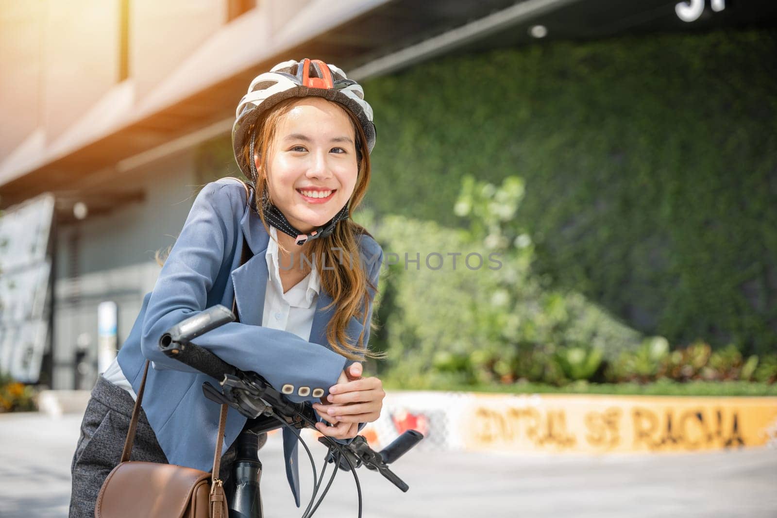 In the city an Asian businesswoman helmeted and in a suit stands with her bicycle ready for a cheerful morning commute to the office. This image combines work and outdoor fun.