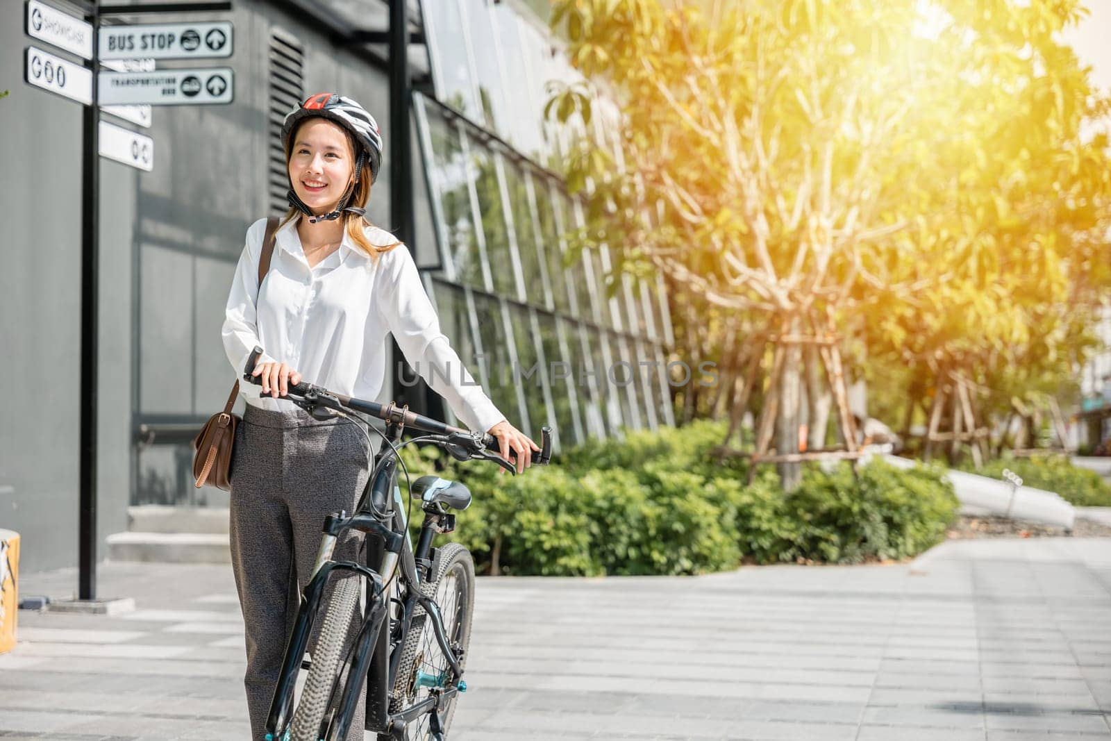 A cheerful Asian businesswoman helmeted and in suit stands with her bicycle symbolizing the modern concept of a joyful business commuter. This image showcases the harmony between work and outdoor fun. by Sorapop