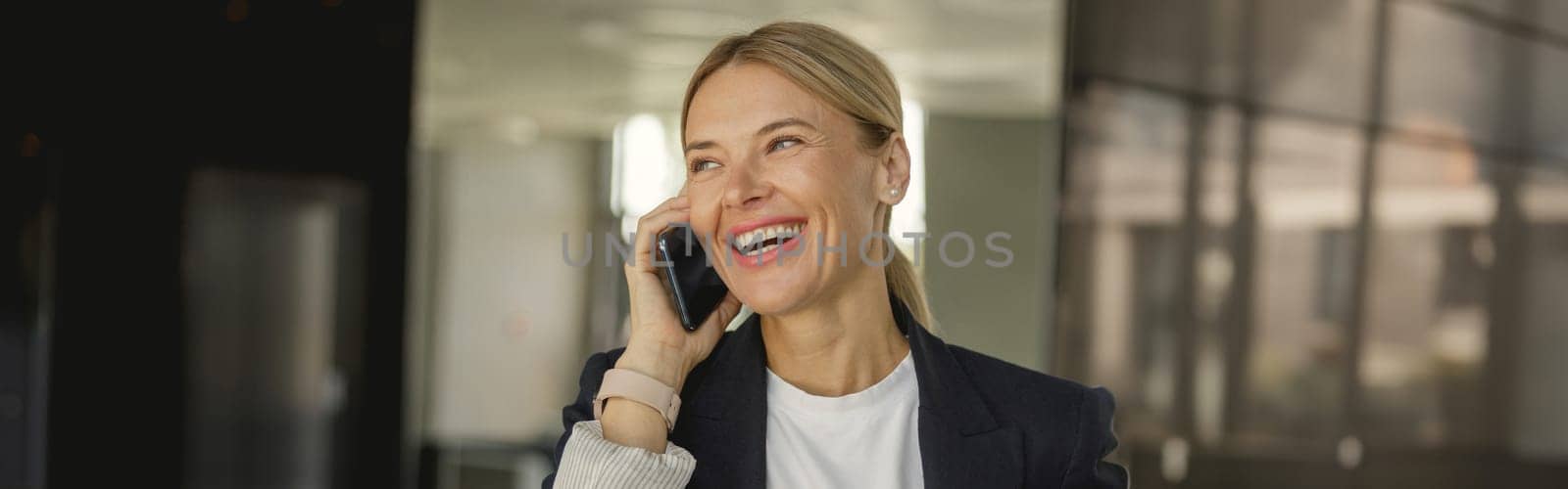 Cheerful businesswoman is talking phone standing on background of modern offfice hall and looks away by Yaroslav_astakhov