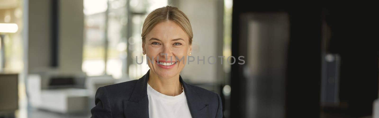 Smiling female entrepreneur with phone standing on background of modern office wall and looks camera