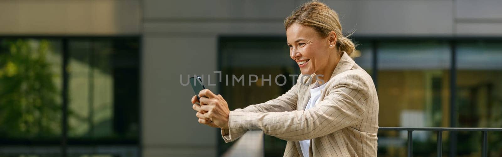 Smiling business woman holding phone while standing on modern office terrace
